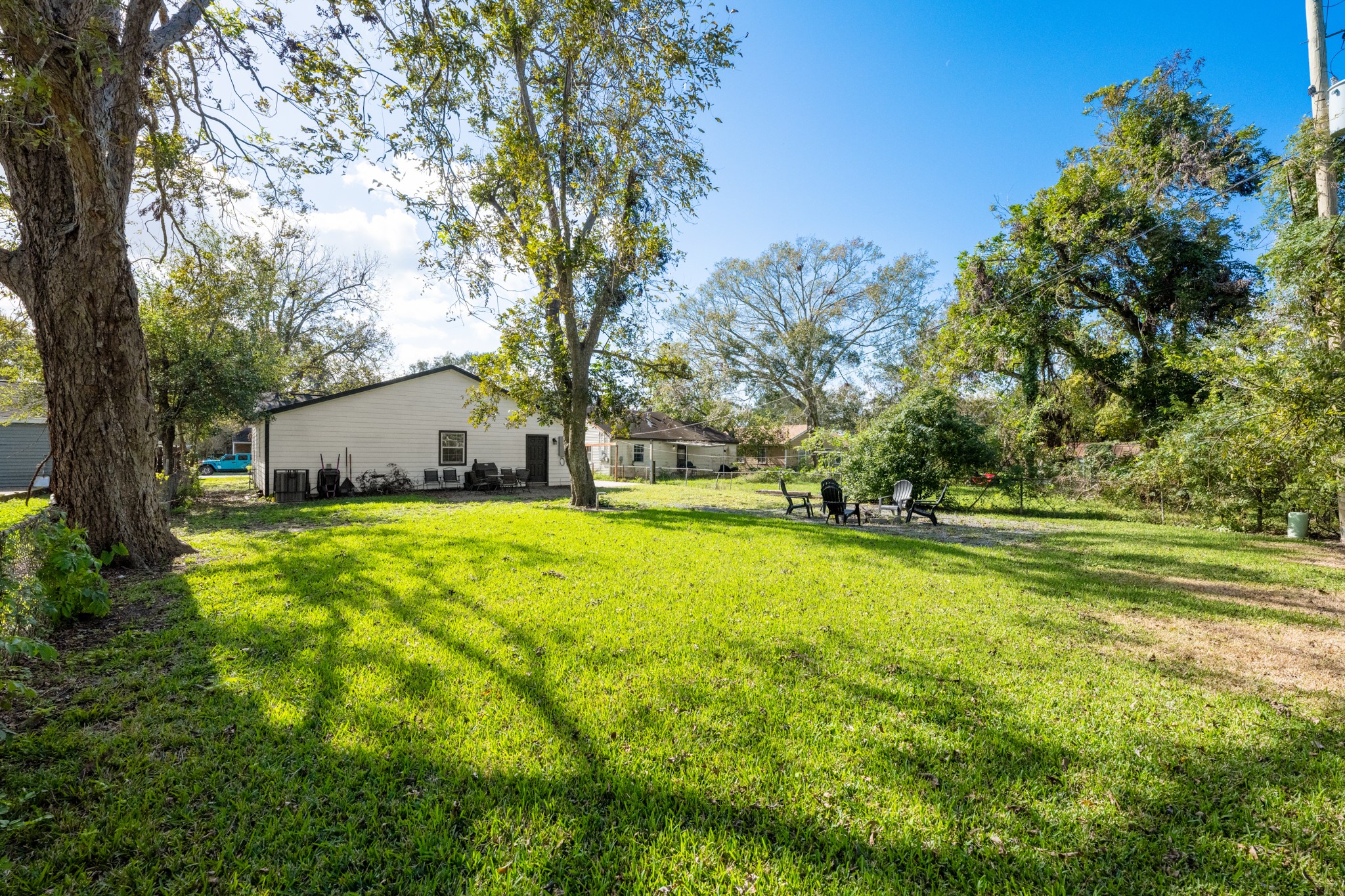 729 Kyle Angleton, TX 77515 - Photo 35 of 36 This photo showcases a spacious backyard with lush green grass and mature trees, providing ample shade and a serene atmosphere. The yard includes a cozy seating area with chairs around a fire pit, ideal for outdoor gatherings. The house in the background has a simple, charming design.