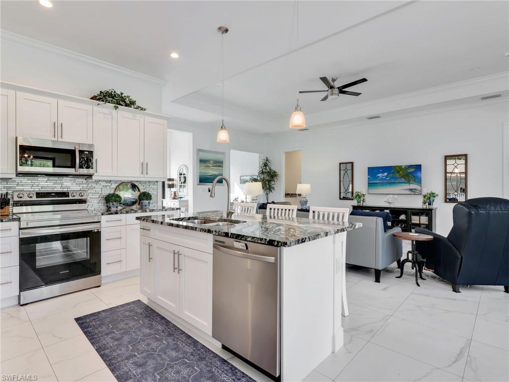 2007 22nd Avenue Northeast Naples, FL 34120 - Photo 12 of 47 Kitchen With White Shaker Cabinetry, Kitchen Island With Double Stainless-Steel Sink, Pendant Lighting, Glass Tile Backsplash, & Glass Tile Backsplash, & Living Room With Tray Ceiling and Ceiling Fan