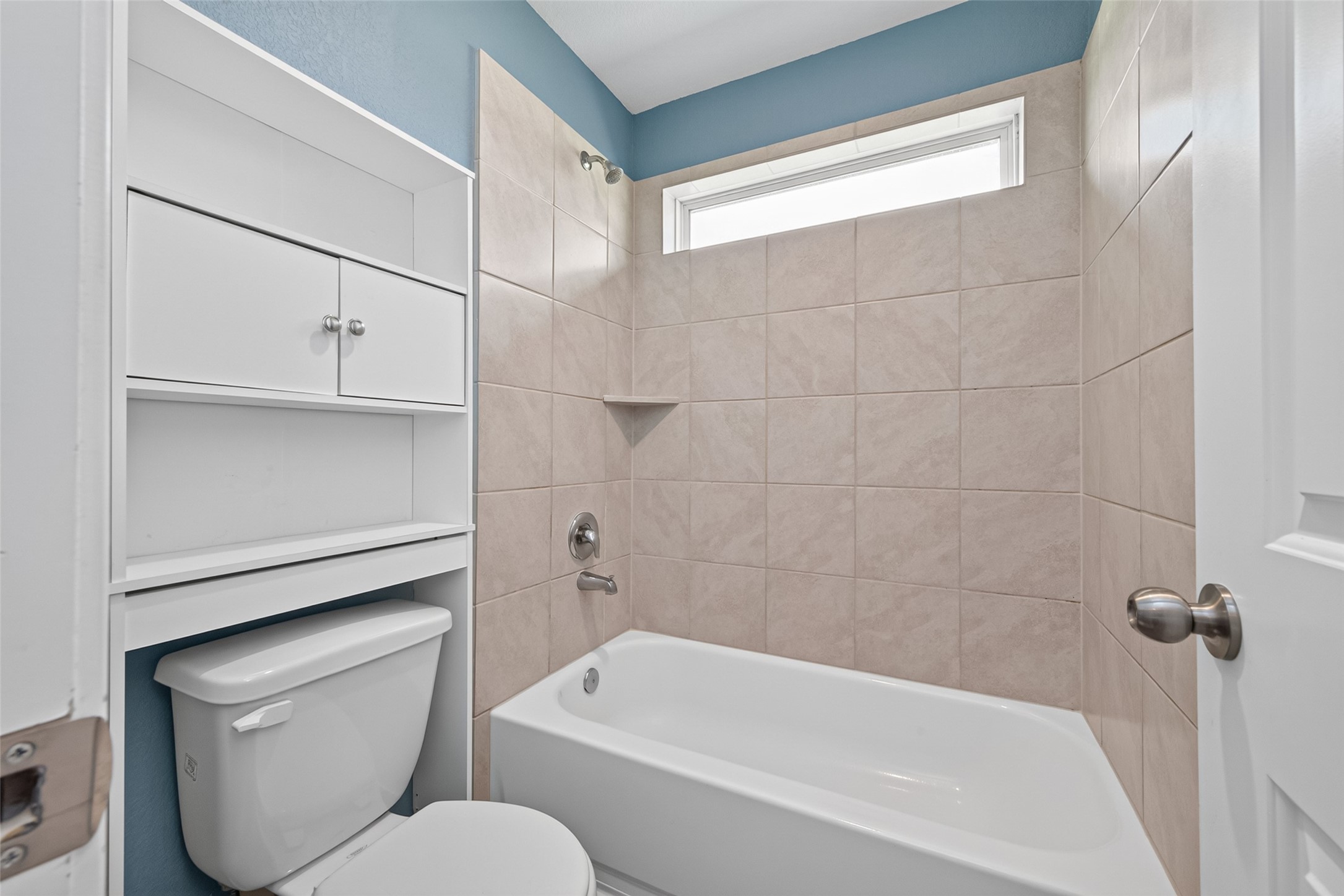 2526 Sturgeon Way Conroe, TX 77384 - Photo 34 of 49 This bathroom features a modern design with beige tiled walls, a white bathtub, and a window for natural light. There is a toilet with built-in shelving above it for extra storage.