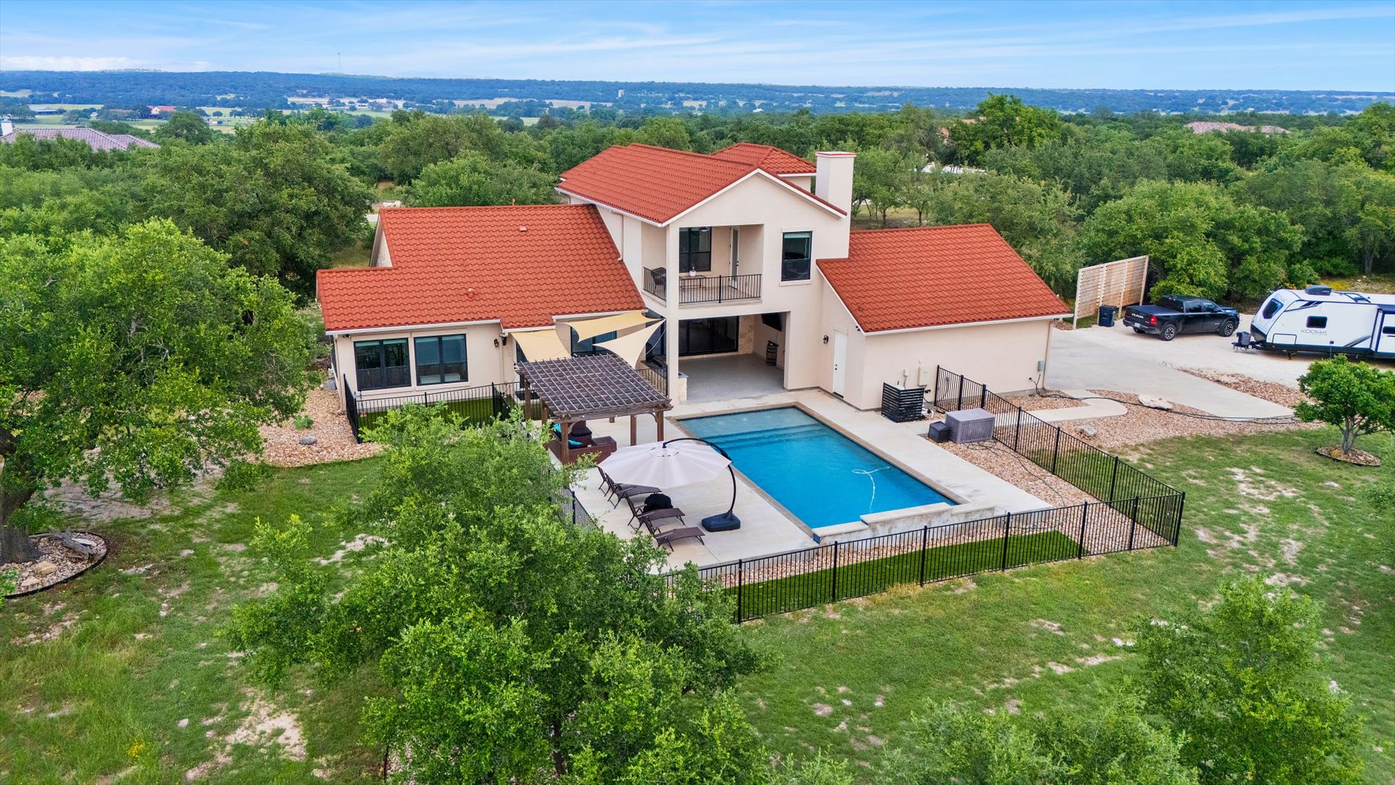 A spacious home with a red tile roof overlooks a verdant landscape, featuring a sparkling pool and inviting outdoor living spaces.