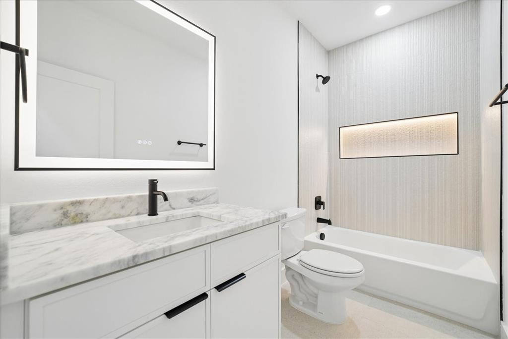 1652 West Wintergreen Road Lancaster, TX 75134 - Photo 19 of 25 Backlit vanity mirror, single basin sink, white cabinetry with dark pull hardware, white tub, and an illuminated shower niche