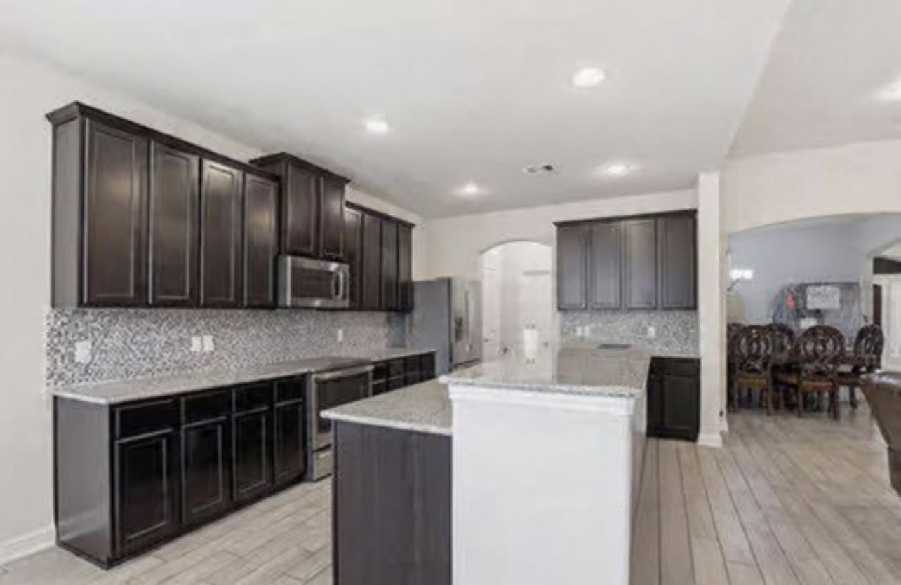 24714 Longwood Forest Drive Spring, TX 77373 - Photo 7 of 21 This modern kitchen features dark wood cabinets, a stylish mosaic tile backsplash, and stainless steel appliances. It offers ample counter space with a central island and opens to a dining area, ideal for entertaining.