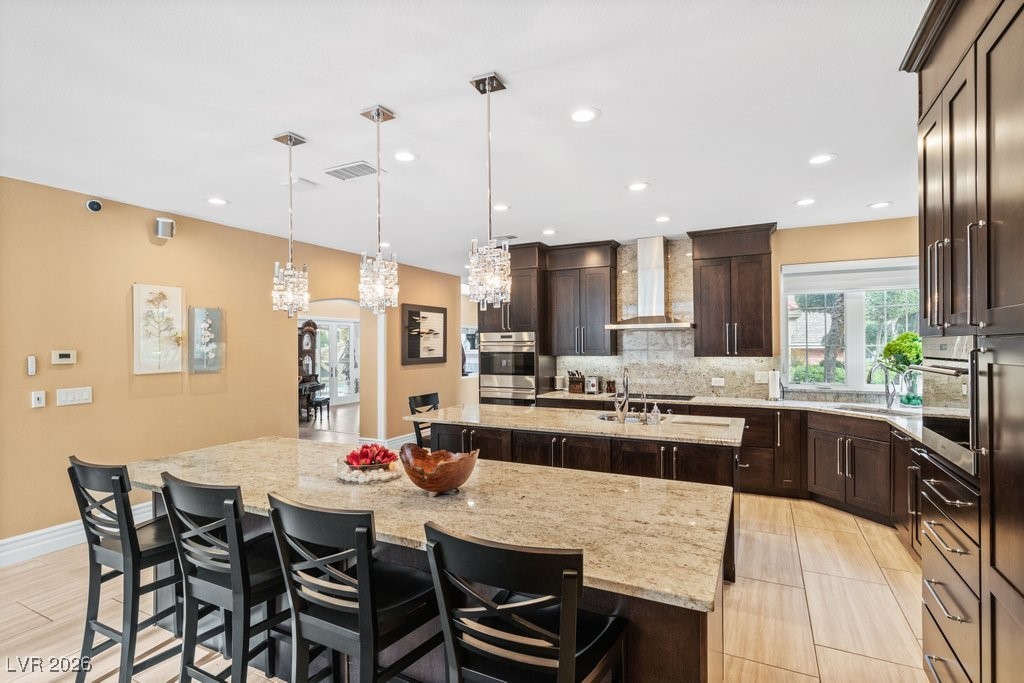 7 Larkside Court Henderson, NV 89014 - Photo 14 of 70 Kitchen with decorative light fixtures, light stone counters, a breakfast bar, dark brown cabinetry, and wall chimney exhaust hood