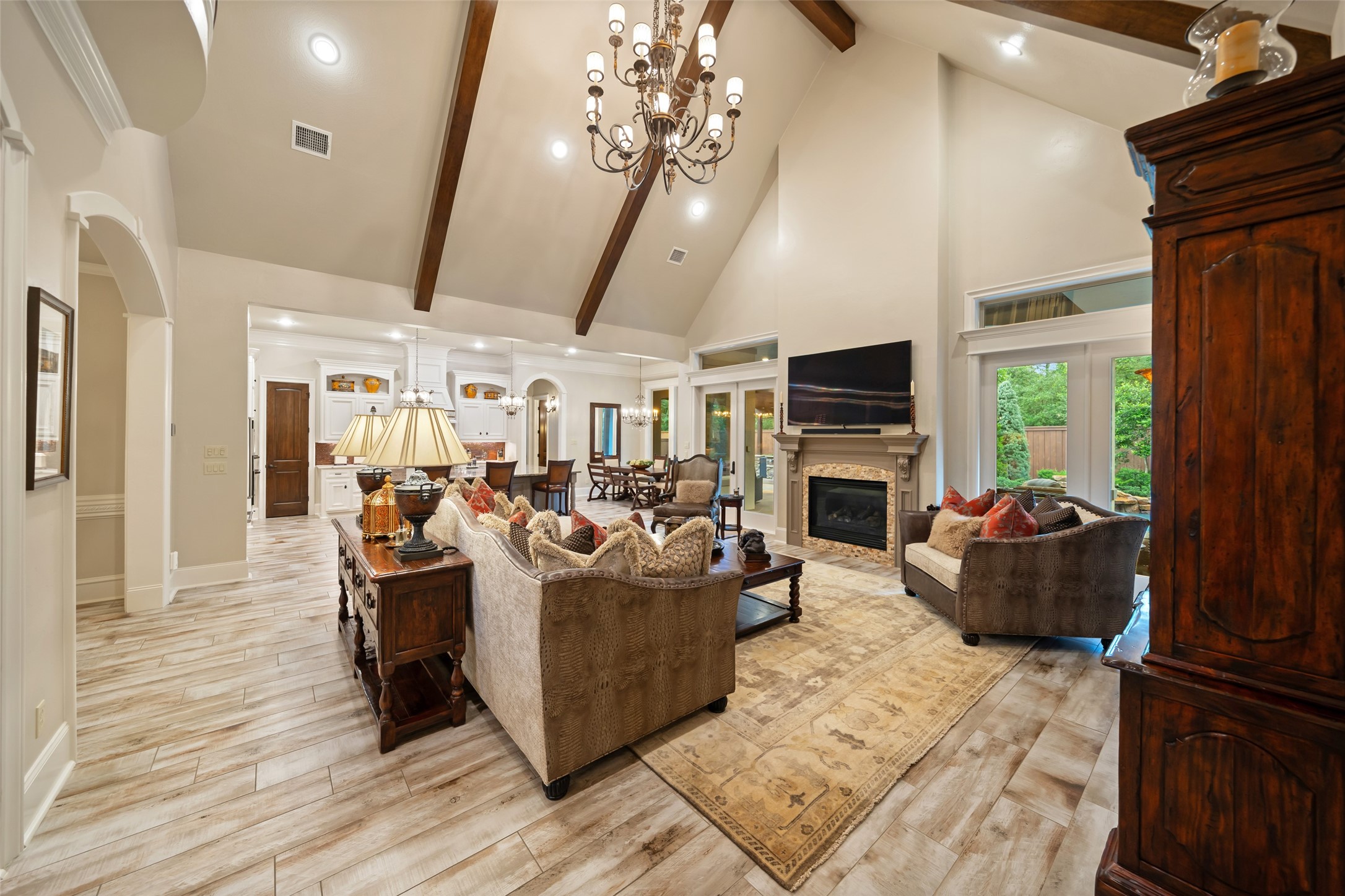 59 East Ambassador Bend Spring, TX 77382 - Photo 7 of 48 Vaulted ceilings, complemented by rich stained wood beams, make this living room truly unforgettable. A grand chandelier serves as a striking centerpiece while expansive French doors and transoms flood the space with natural light, enhancing the room’s warmth and openness. The combination of architectural details and a bright, airy ambiance creates an inviting yet dramatic setting, perfect for both relaxation and entertainment.