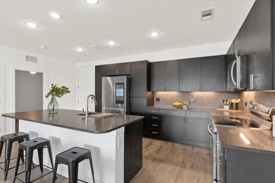 4801 Springdale Road, Unit 1107 Austin, TX 78723 - Photo 6 of 40 Kitchen with stainless steel appliances, a kitchen island with sink, dark cabinetry, light wood-style floors, and a kitchen bar