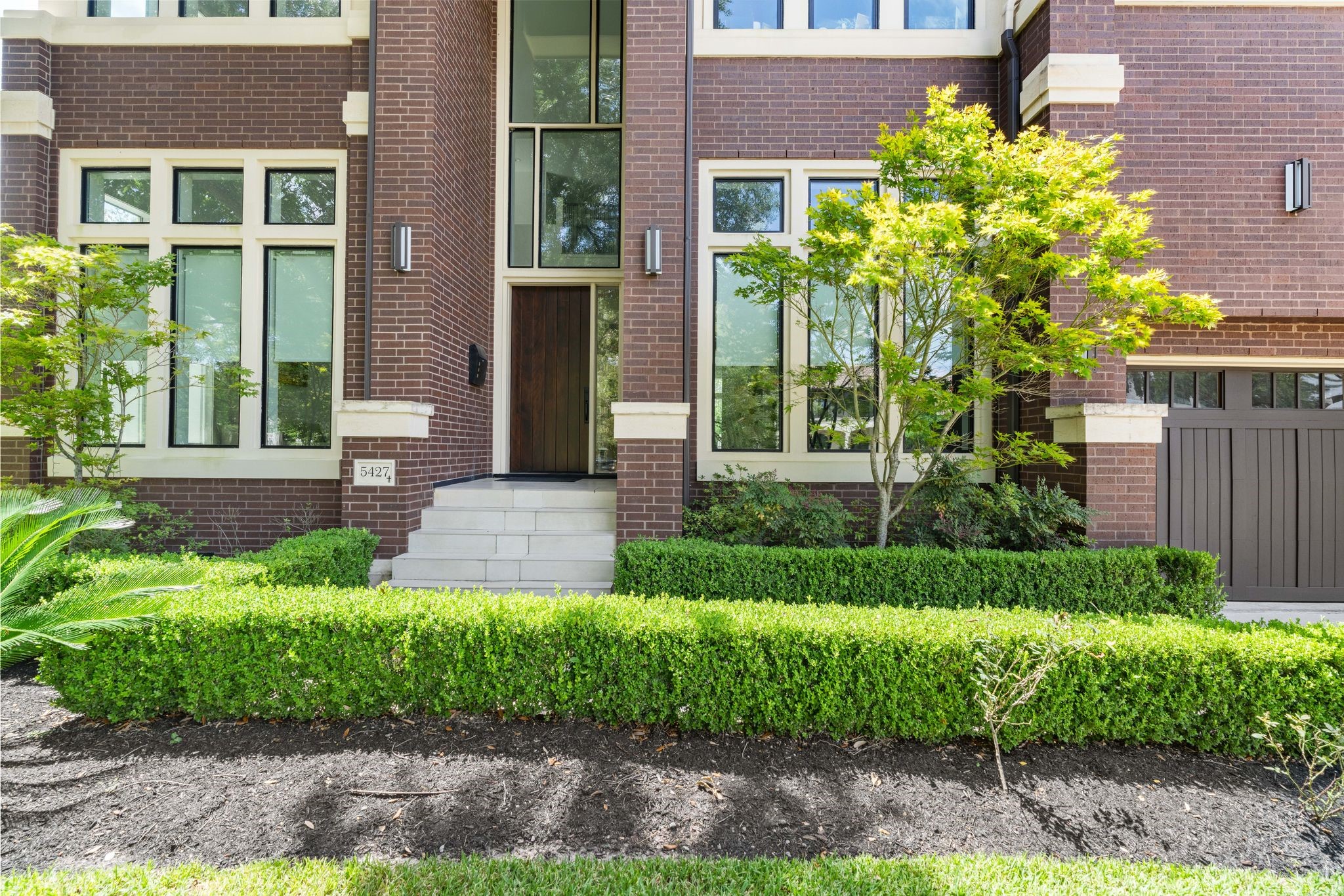 5427 Maple Street Houston, TX 77096 - Photo 6 of 46 A closer look at the front door reveals a solid wood entry set within a dramatic glass surround, flanked by brick columns with stone caps. A short run of stone steps leads up through a neatly trimmed boxwood hedge, with a young tree adding color beside the door.