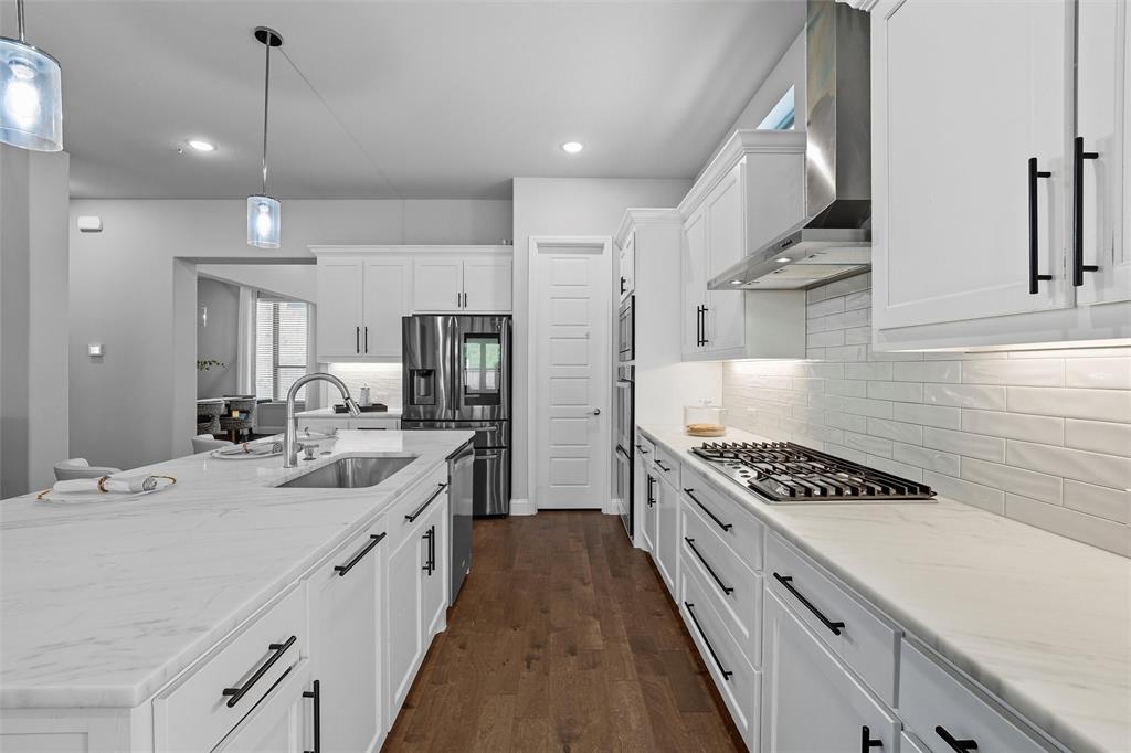8492 Chapote Road Frisco, TX 75035 - Photo 19 of 40 Kitchen features pendant lights over a large center island. Stainless steel appliances including double ovens, microwave, and gas cooktop with stainless steel vent hood. White cabinetry with modern hardware and tile backsplash. Large island with quartz countertop and undermount sink. Wide-plank hardwood flooring extending through the space.