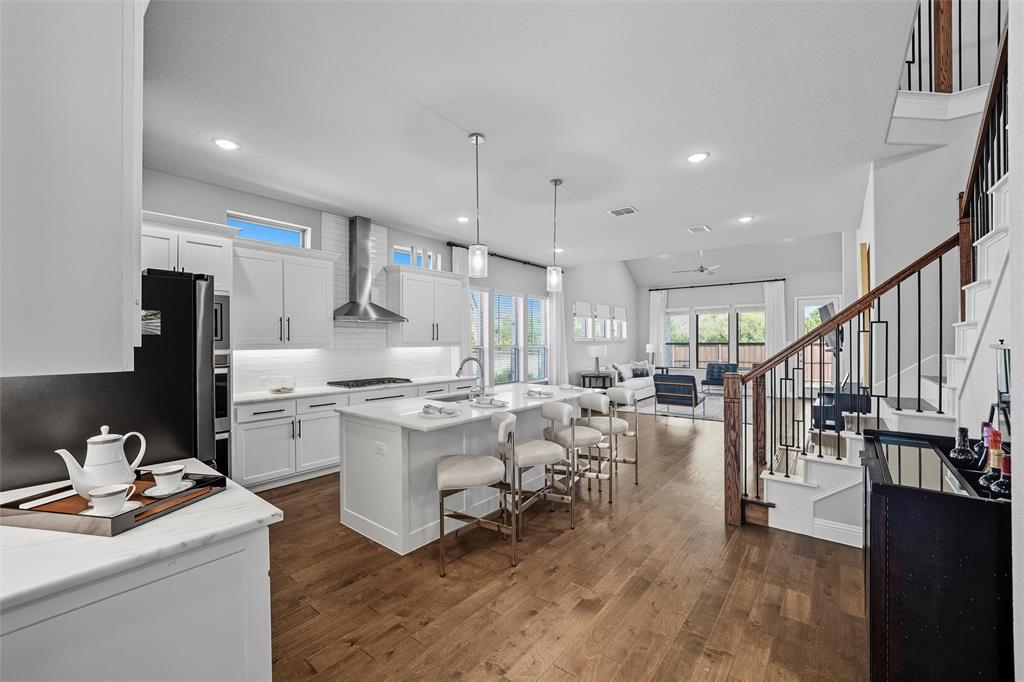 8492 Chapote Road Frisco, TX 75035 - Photo 21 of 40 Kitchen features pendant lights over a large center island. Stainless steel appliances including double ovens, microwave, and gas cooktop with stainless steel vent hood. White cabinetry with modern hardware and tile backsplash. Large island with quartz countertop and undermount sink. Wide-plank hardwood flooring extending through the space.