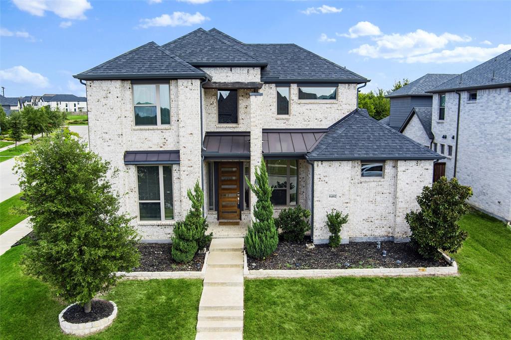 8492 Chapote Road Frisco, TX 75035 - Photo 30 of 40 Front Exterior features light-colored brick façade with upper and lower windows framed in dark trim. Covered entry with wood beam detail above the doorway. Modern wood front door with horizontal glass inserts. Evergreen shrubs and manicured landscaping in stone-edged beds. Wide concrete steps and walkway leading to the entrance across a green lawn.