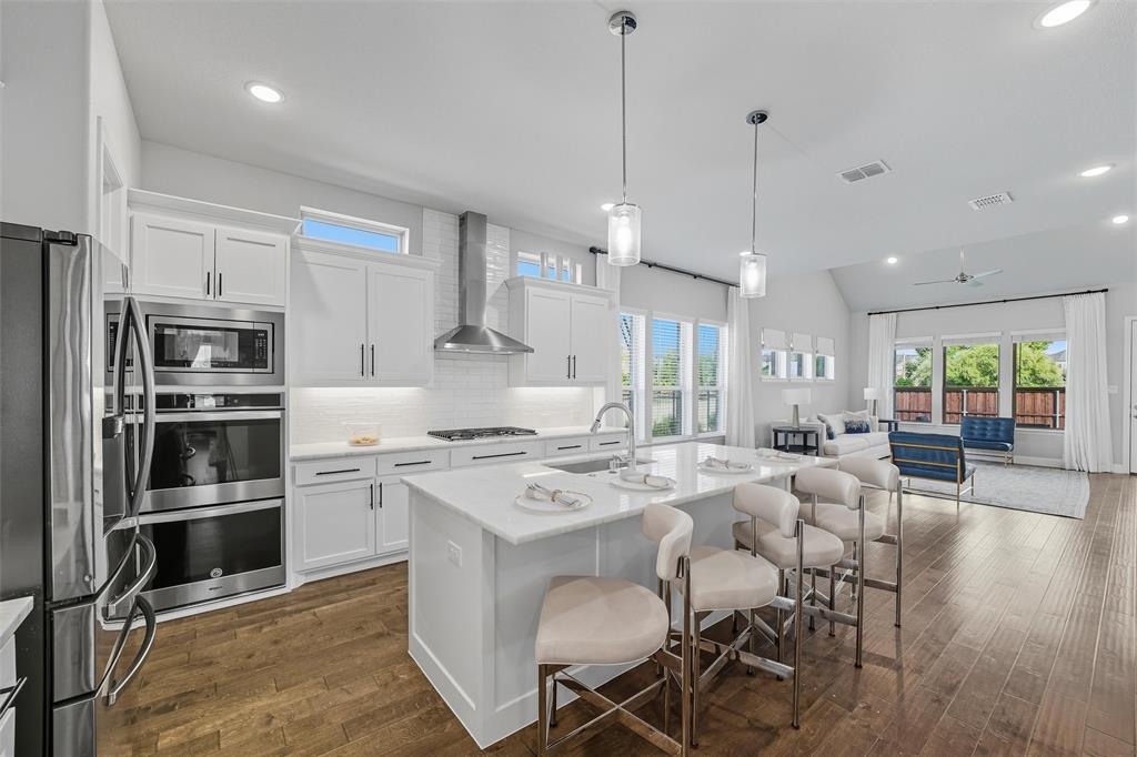 8492 Chapote Road Frisco, TX 75035 - Photo 6 of 40 Kitchen features pendant lights over a large center island. Stainless steel appliances including double ovens, microwave, and gas cooktop with stainless steel vent hood. White cabinetry with modern hardware and tile backsplash. Large island with quartz countertop and undermount sink. Wide-plank hardwood flooring extending through the space.