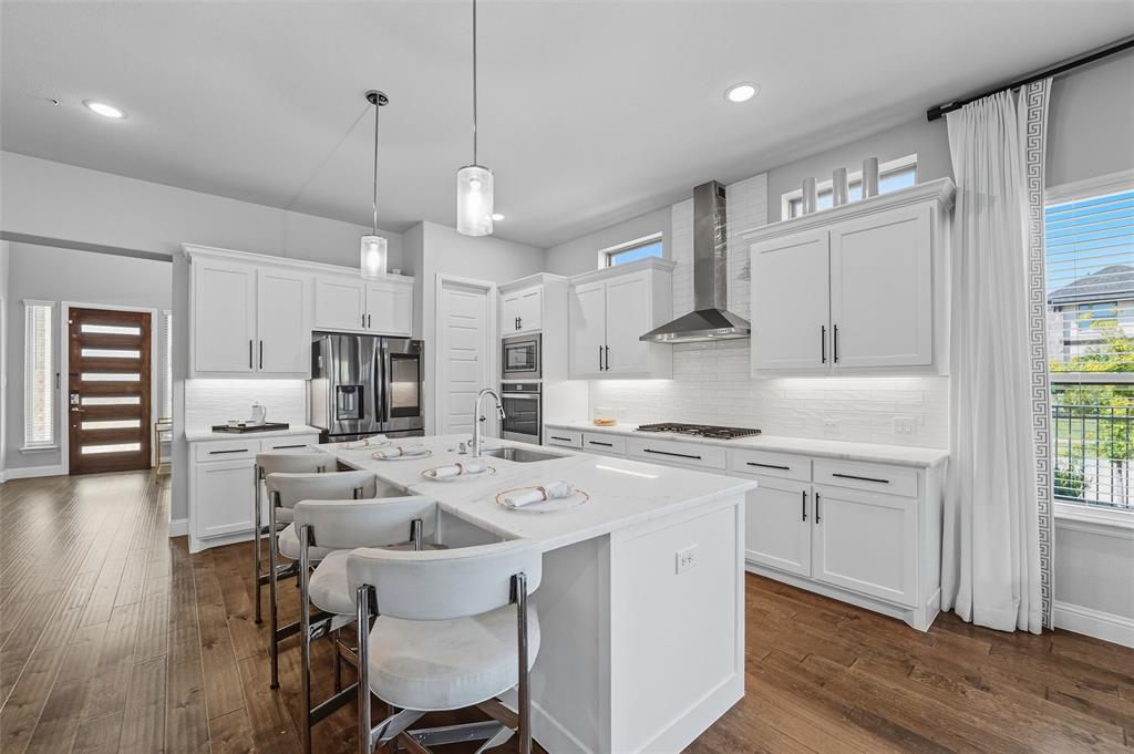 8492 Chapote Road Frisco, TX 75035 - Photo 7 of 40 Kitchen features pendant lights over a large center island. Stainless steel appliances including double ovens, microwave, and gas cooktop with stainless steel vent hood. White cabinetry with modern hardware and tile backsplash. Large island with quartz countertop and undermount sink. Wide-plank hardwood flooring extending through the space.