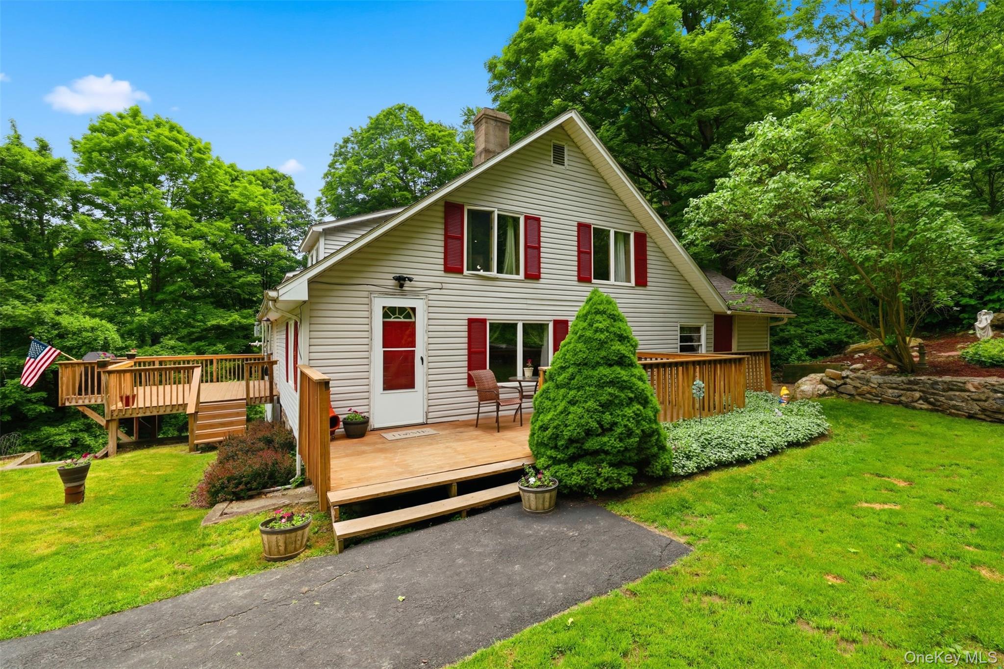 Gable-front home featuring light-colored siding, red window shutters, and two wooden decks with railings surrounded by green grass and mature trees