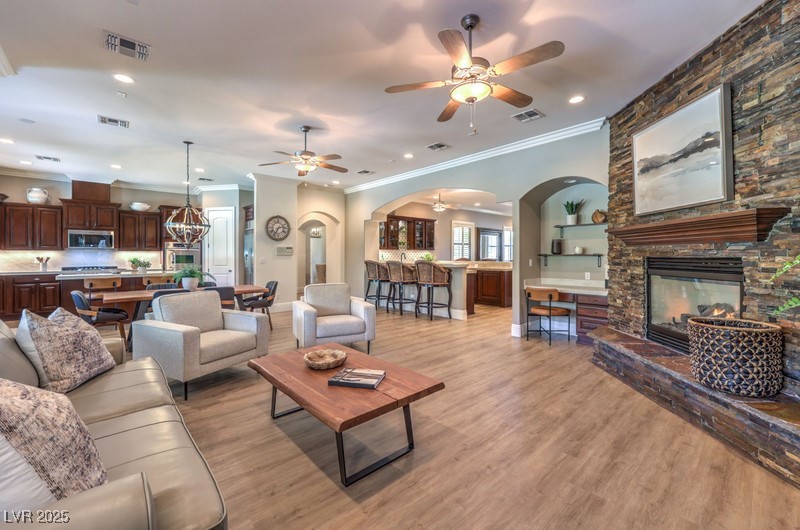 9135 Hickam Avenue Las Vegas, NV 89129 - Photo 15 of 75 Living room featuring a stone fireplace, arched walkways, ornamental molding, a ceiling fan, and light wood-type flooring