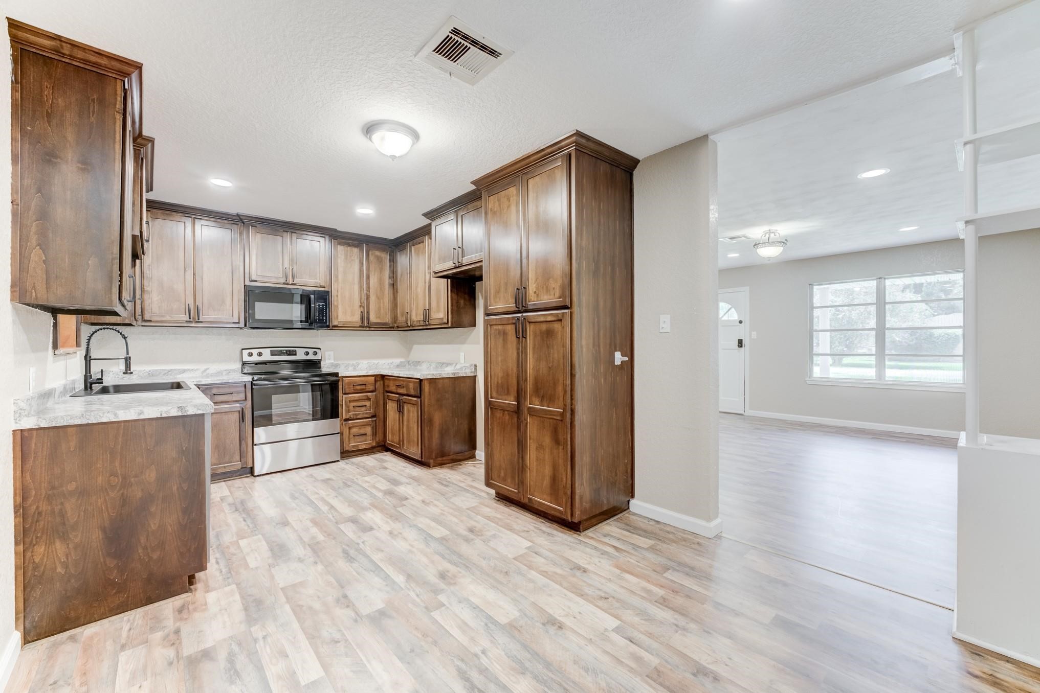 305 Yaupon Street Sweeny, TX 77480 - Photo 17 of 22 This photo showcases a spacious, open-concept kitchen and living area. The kitchen features modern stainless steel appliances, ample wooden cabinetry, and marble-look countertops. The adjacent living space is bright, with large windows allowing natural light to flow in, and light wood flooring throughout enhances the airy feel.