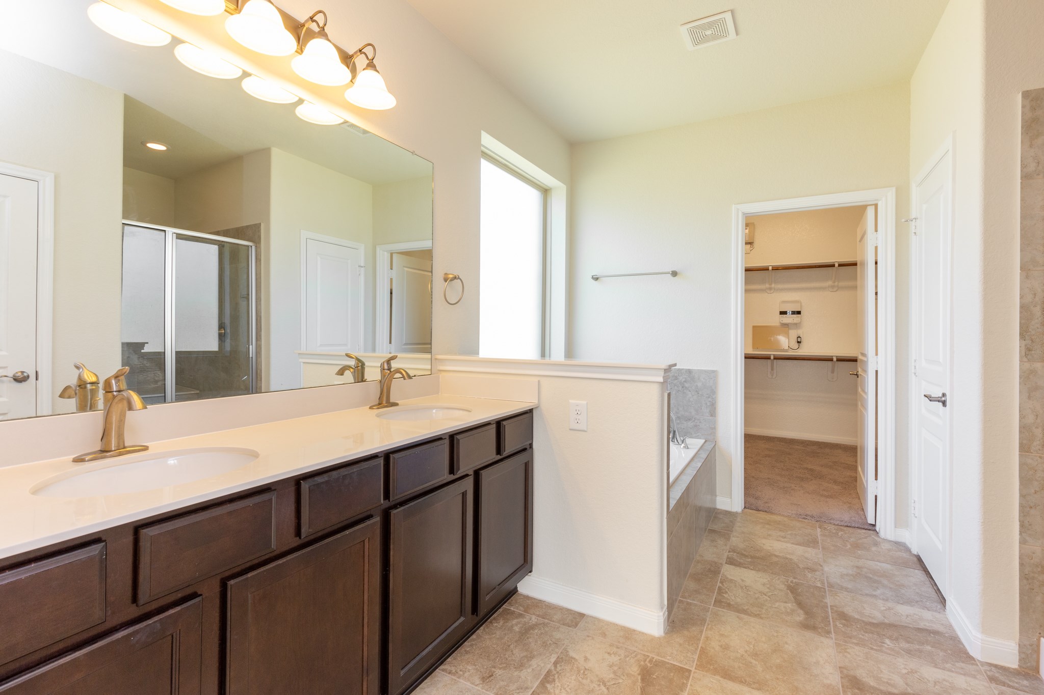 10 Log House Court Tomball, TX 77375 - Photo 32 of 50 This bathroom features a spacious layout with dual sinks set in a long vanity with dark wood cabinets. It includes a large mirror, a glass-enclosed shower, and a separate bathtub. Natural light streams in through a generous window, and there's direct access to a walk-in closet. The neutral tile flooring complements the elegant fixtures.