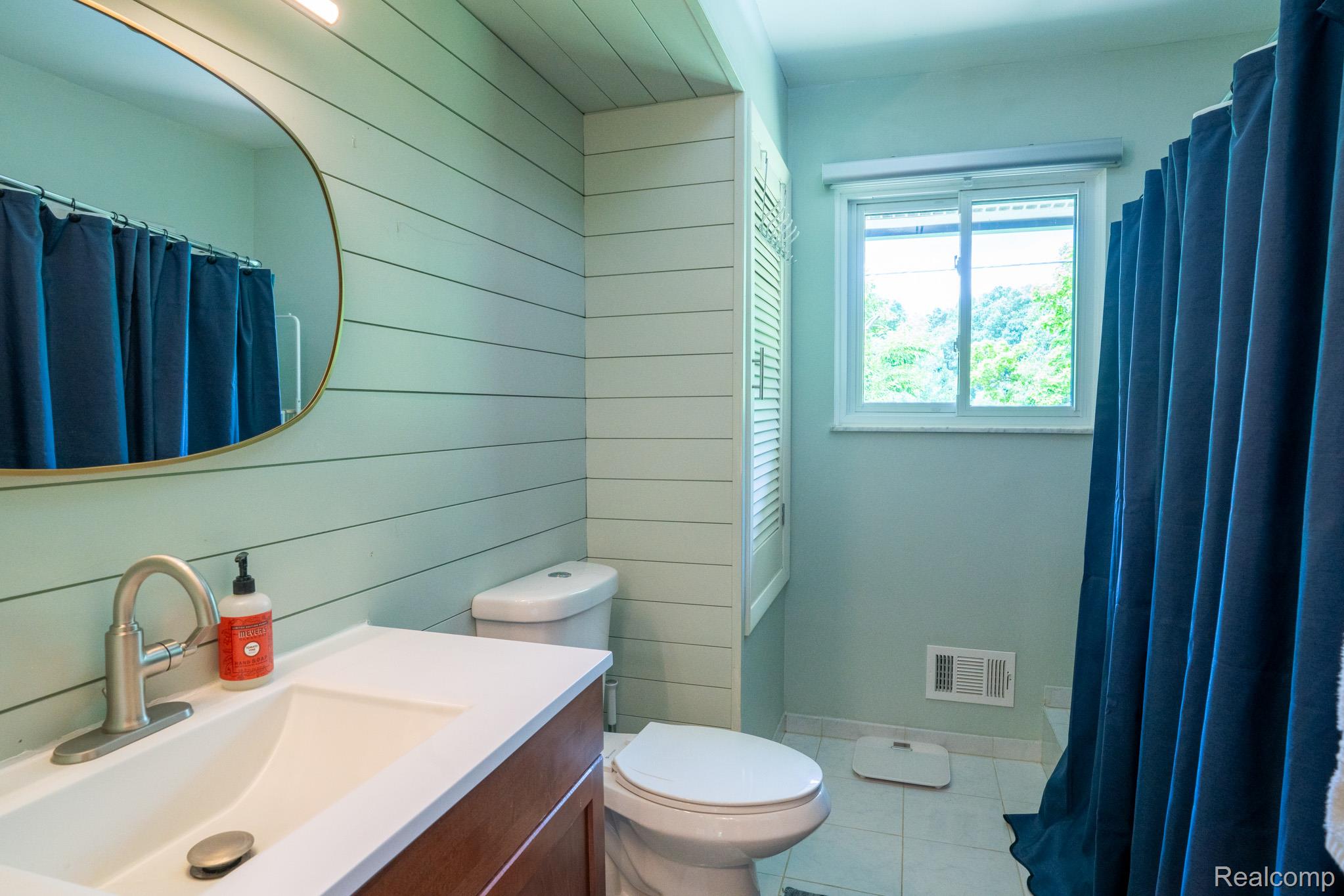 7333 Toma Road Dexter, MI 48130 - Photo 26 of 45 Bathroom featuring shiplap wall paneling, a single vanity with a white countertop and undermount sink, a wall-mounted oval mirror, a white toilet, and a window providing natural illumination