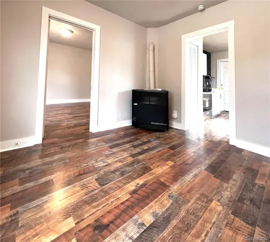 an empty room with wooden floor and way to kitchen
