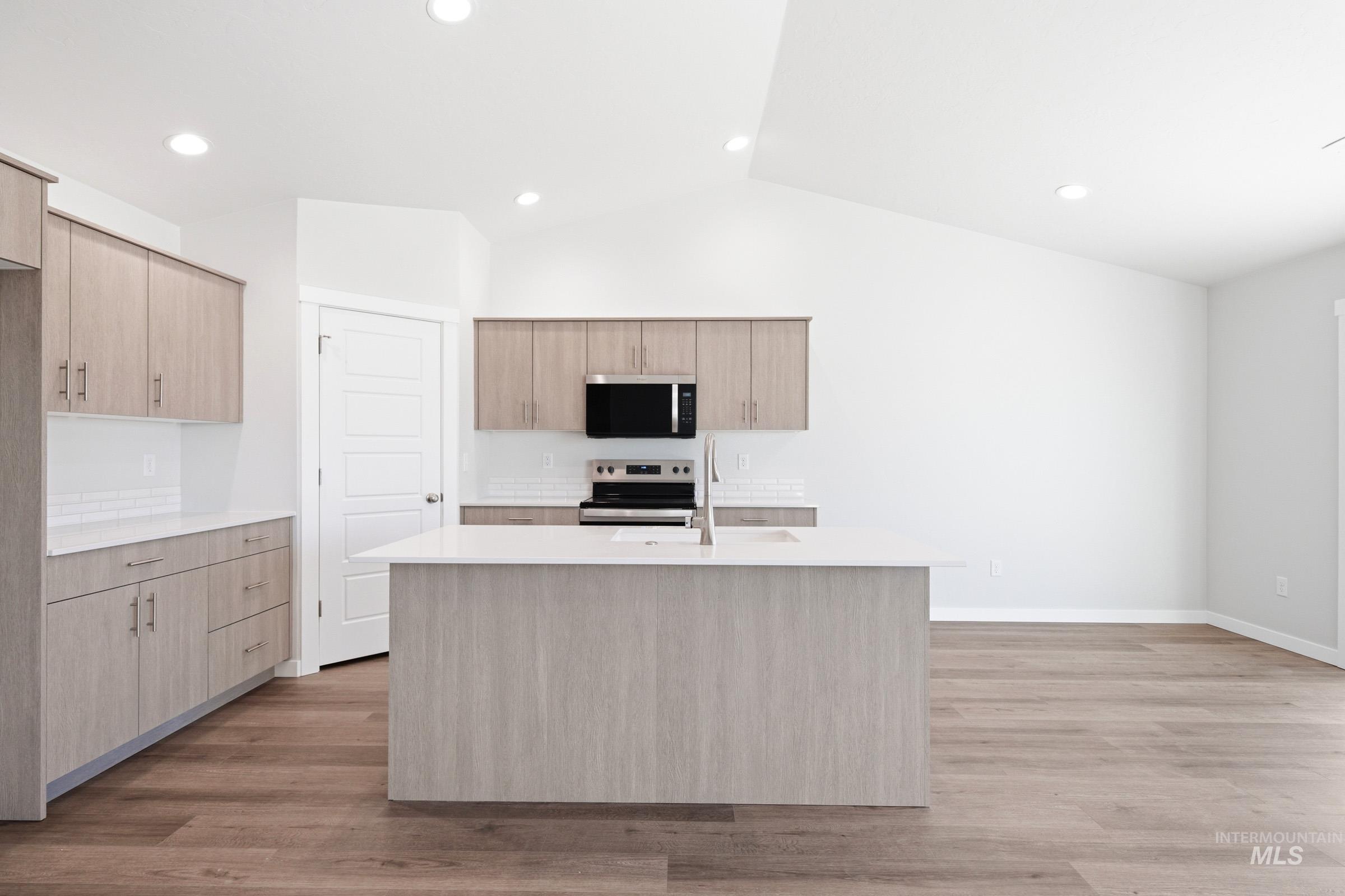 12877 Decatur Street Nampa, ID 83651 - Photo 3 of 22 Modern kitchen featuring a central island with a sink and a stainless steel faucet, wood-finish flooring, light wood-look cabinetry, white countertops, and a white subway tile backsplash