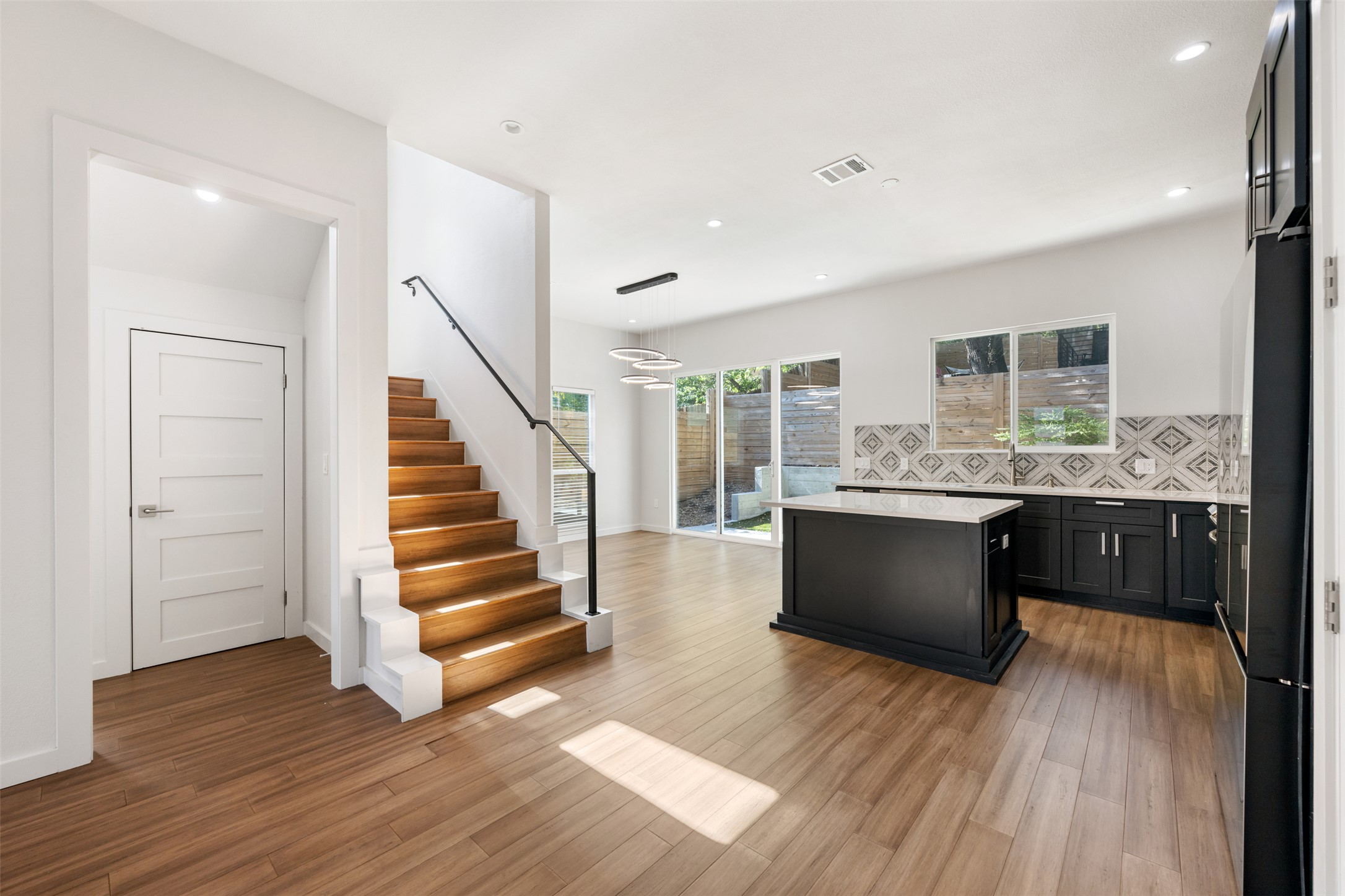 3010 Webberville Road, Unit 1 Austin, TX 78702 - Photo 3 of 38 Open-concept living space with wood-finish flooring, a contemporary kitchen featuring dark cabinetry and a patterned tile backsplash, and a staircase with a black metal railing