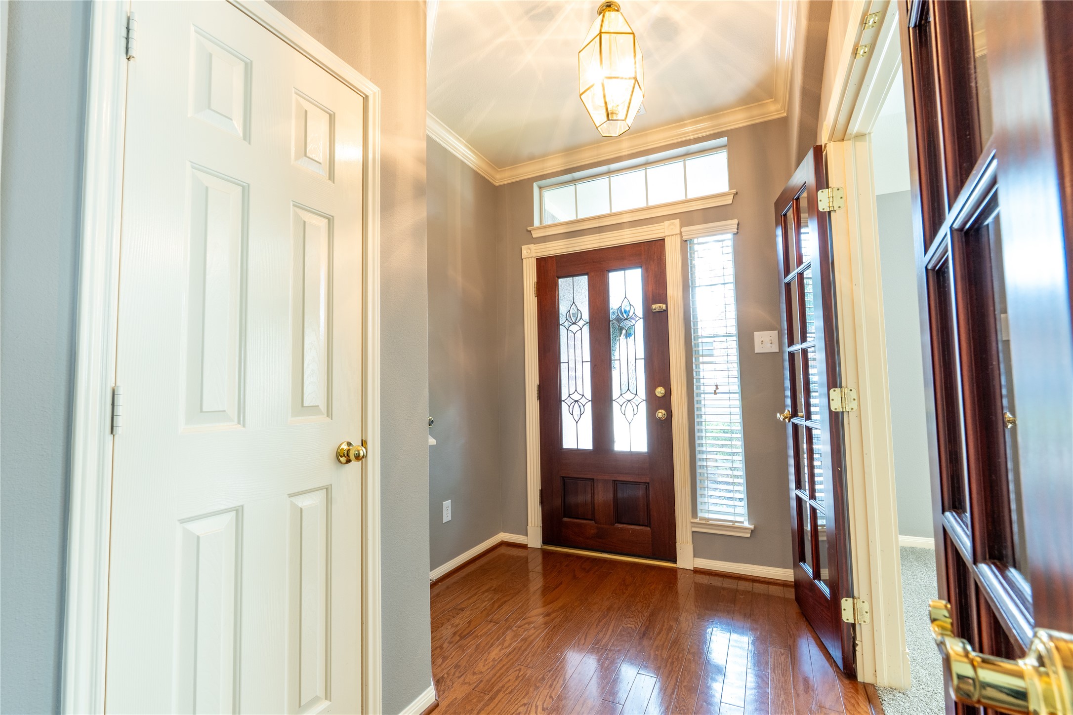 10619 Crownsedge Drive Spring, TX 77379 - Photo 28 of 38 This photo shows a welcoming entryway with a wooden front door featuring decorative glass panels. The space has hardwood floors, crown molding, and ample natural light from tall side windows. A stylish light fixture adds elegance to the area.