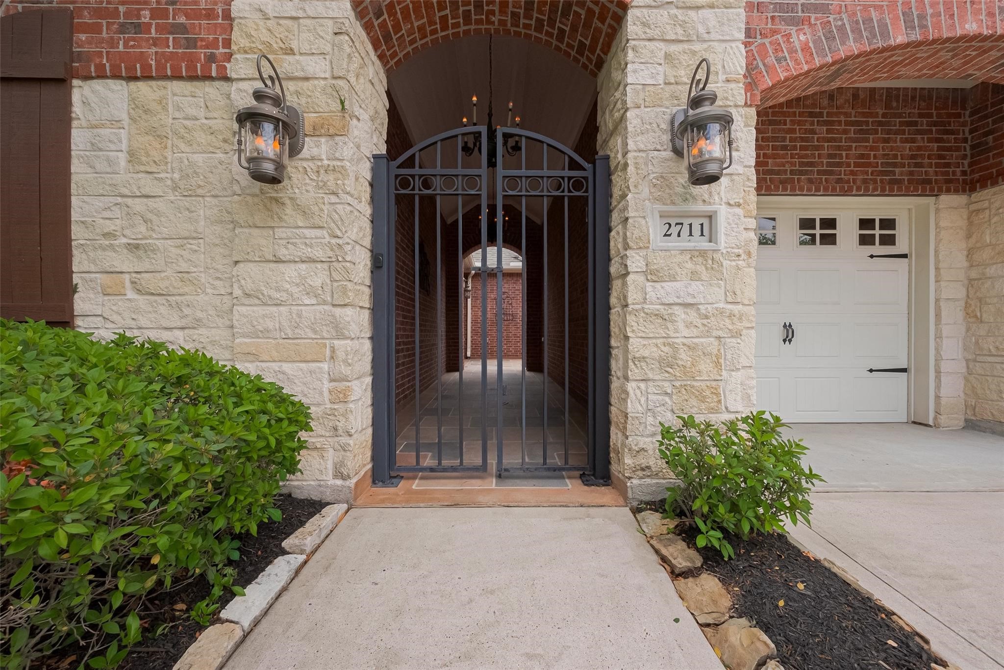 2711 King Point View Lane Spring, TX 77388 - Photo 3 of 48 This inviting entrance features a charming wrought iron gate, framed by elegant stonework and brick, with stylish lanterns providing a warm welcome. The pathway leads to a covered entryway, adjacent to a garage with classic white doors.