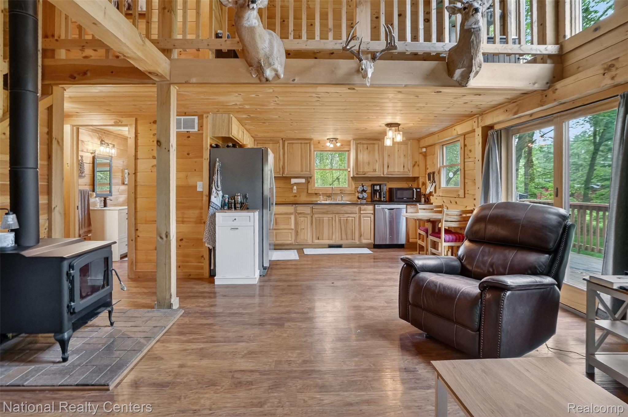 7782 Golf Club Road Howell, MI 48843 - Photo 7 of 27 Open-concept living area with wood-finish flooring, a black wood-burning stove on a tile hearth, and abundant natural wood paneling