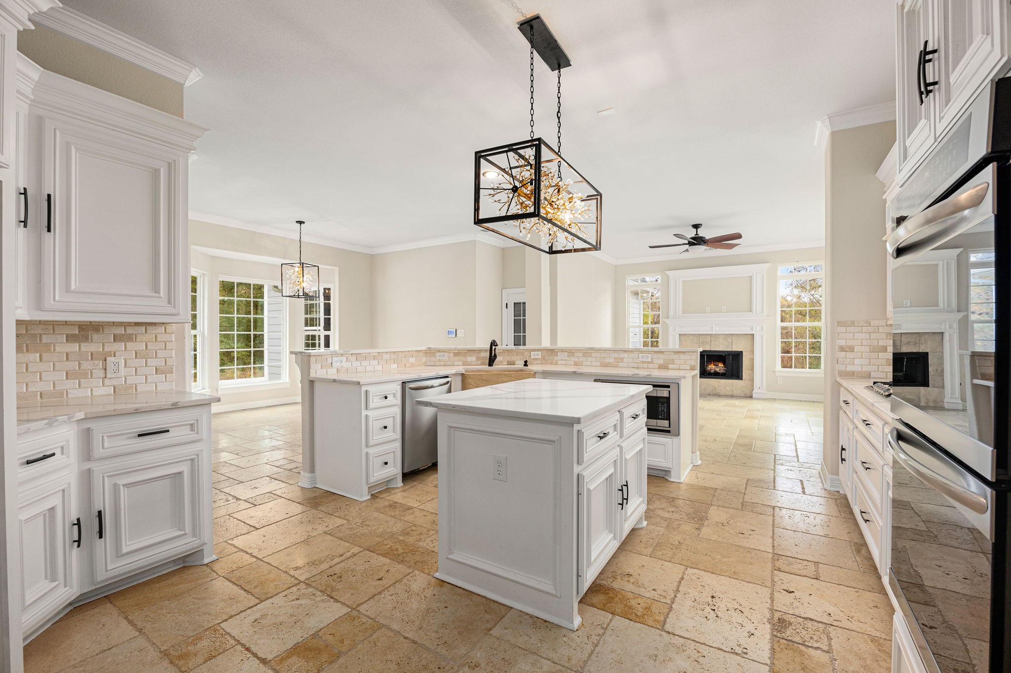 14882 Gingham Road Montgomery, TX 77356 - Photo 15 of 41 This photo showcases a spacious, bright kitchen with elegant white cabinetry, a large central island, and modern pendant lighting. The open layout flows into a cozy living area featuring a fireplace and large windows for natural light. The tiled floor adds a warm, inviting touch.