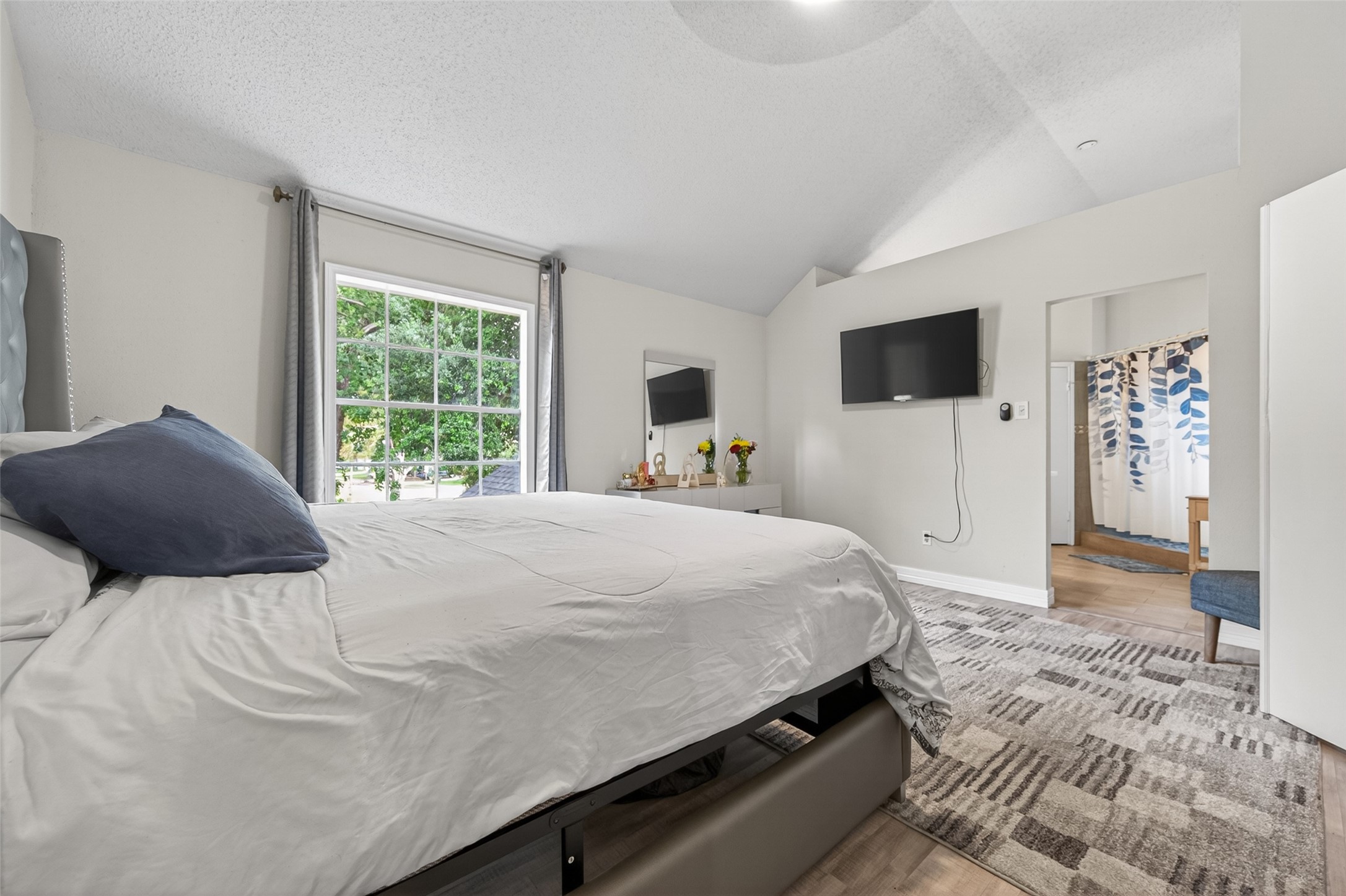 18446 North Willow Bluff Road Katy, TX 77449 - Photo 19 of 33 This bedroom features a large window with lush green views, a cozy bed, carpeted flooring, and a wall-mounted TV. The space is bright and airy, with an adjoining bathroom visible through an open doorway.