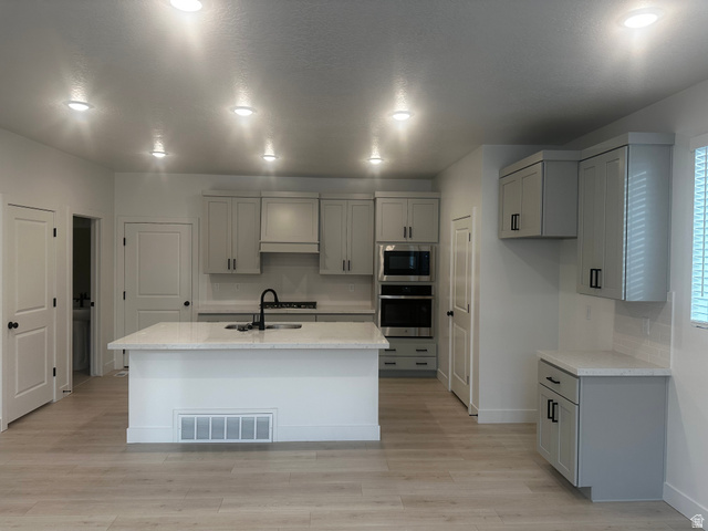 15 South Lunar Way Mapleton, UT 84664 - Photo 3 of 13 Kitchen with gray cabinetry, light wood-style flooring, stainless steel appliances, light stone countertops, and a kitchen island with sink