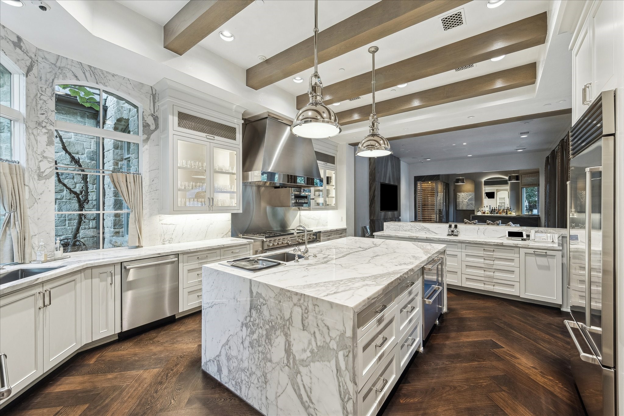 220 Merrie Way Lane Houston, TX 77024 - Photo 10 of 50 This view highlights the kitchen’s thoughtful craftsmanship—from the marble countertops and custom stained beam ceiling to glass-front display cabinetry with LED lighting. An oversized waterfall island with a secondary vegetable sink anchors the space, while windows brighten the Segreto-plastered walls. Smart features like an appliance garage, built-in spice and knife storage, and soft-close cabinetry complete the kitchen.