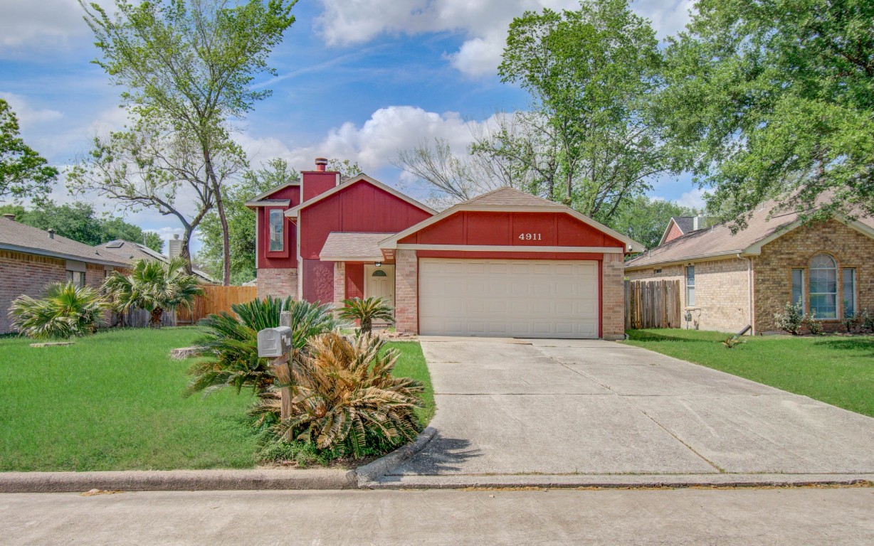 Charming two-story red house with a large driveway and attached garage. The front yard features lush greenery with small palms and a well-maintained lawn, offering a welcoming curb appeal.