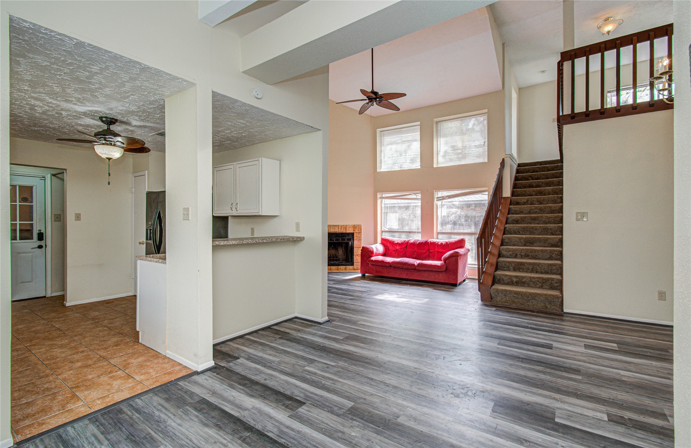 4911 Tealgate Drive Spring, TX 77373 - Photo 14 of 41 This photo showcases an open-concept living area with high ceilings and large windows that allow natural light to flood the space. It features modern gray flooring, a cozy fireplace, and a staircase leading to the upper level. Adjacent is a kitchen area with tiled flooring and a ceiling fan.
