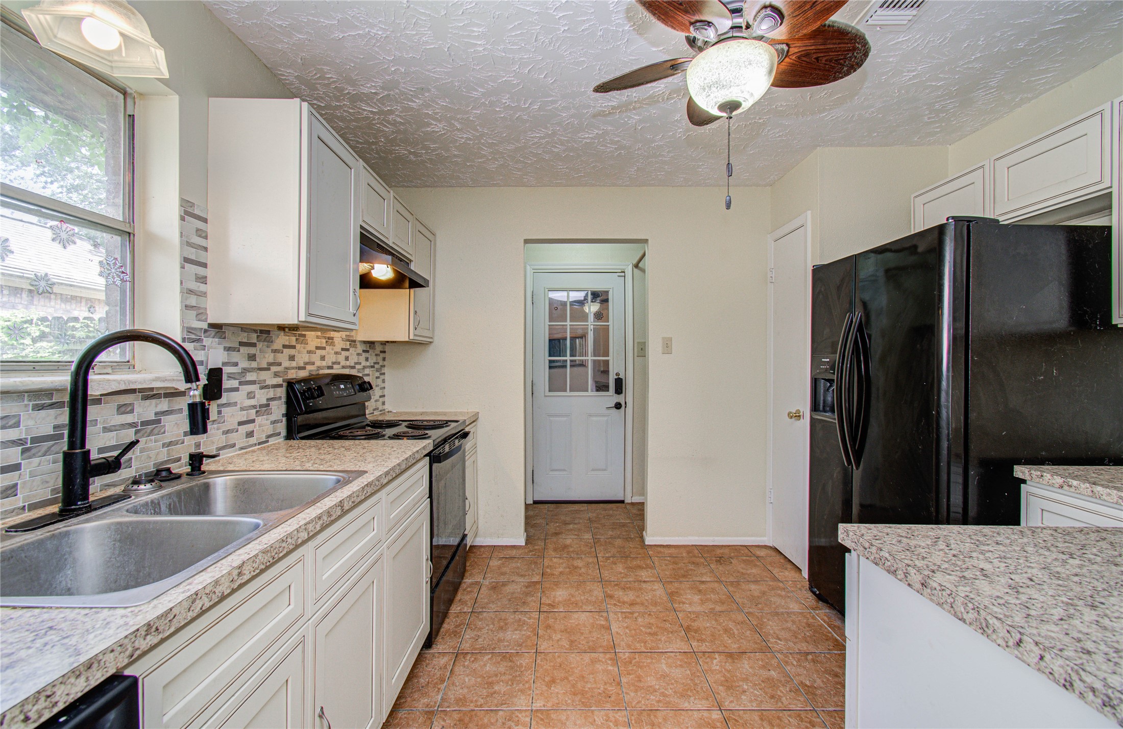 4911 Tealgate Drive Spring, TX 77373 - Photo 16 of 41 This kitchen features modern tile flooring, granite countertops, and a stylish backsplash. There's ample cabinetry, a double sink, black appliances, and a ceiling fan, with a door leading outside.