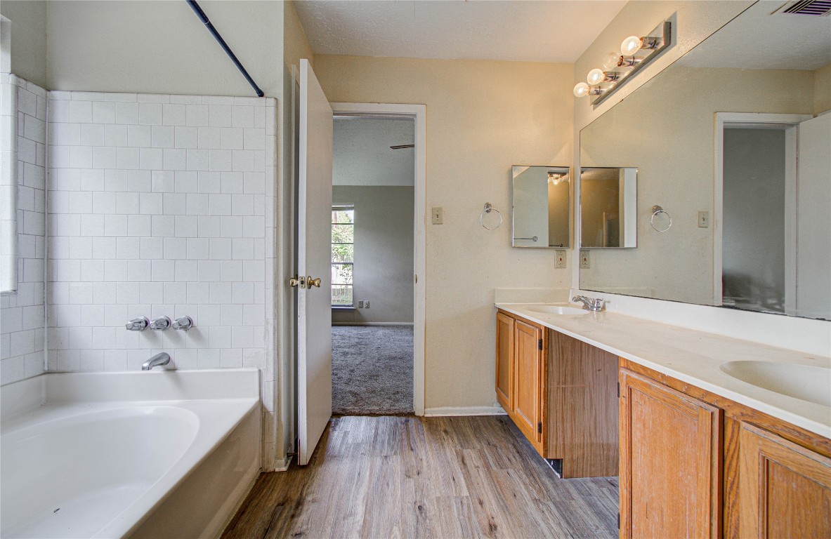 4911 Tealgate Drive Spring, TX 77373 - Photo 27 of 41 This bathroom features a white bathtub with tile surround, dual sinks with wooden cabinetry, a large mirror, and wood-look flooring. The space connects to a carpeted room with natural light.