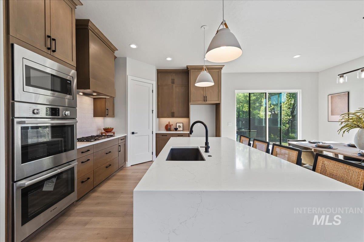 985 West Malbar Street Meridian, ID 83646 - Photo 11 of 39 Modern kitchen featuring a large center island with a black sink and faucet, stainless steel double ovens and microwave, a gas cooktop, wood-finish cabinetry, and light wood-finish flooring