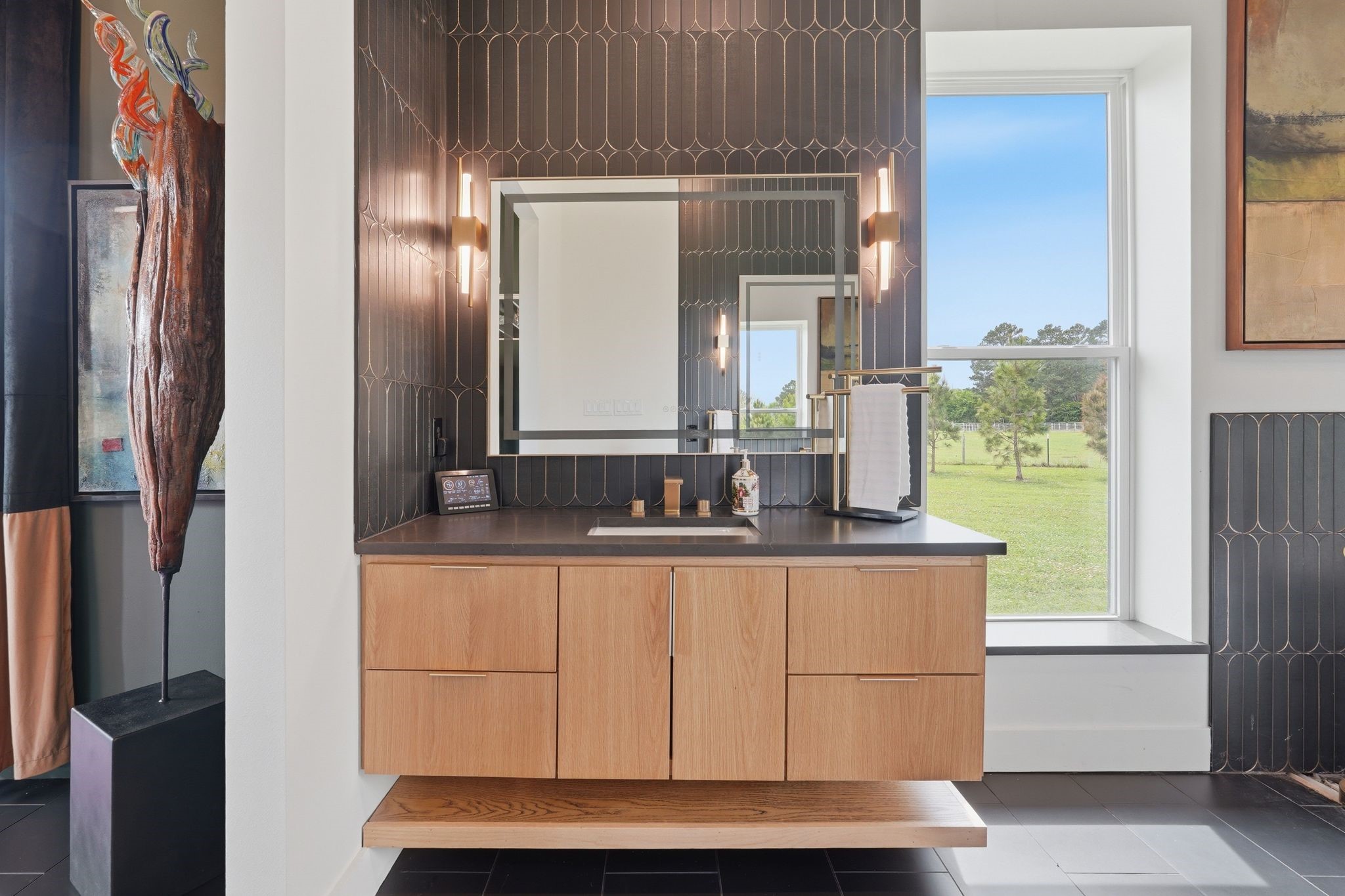 2743 Bobville Road Montgomery, TX 77316 - Photo 30 of 50 This modern bathroom features a sleek, floating wooden vanity with a dark countertop, set against stylish dark tile walls. It includes a large, well-lit mirror and a window offering a scenic outdoor view.