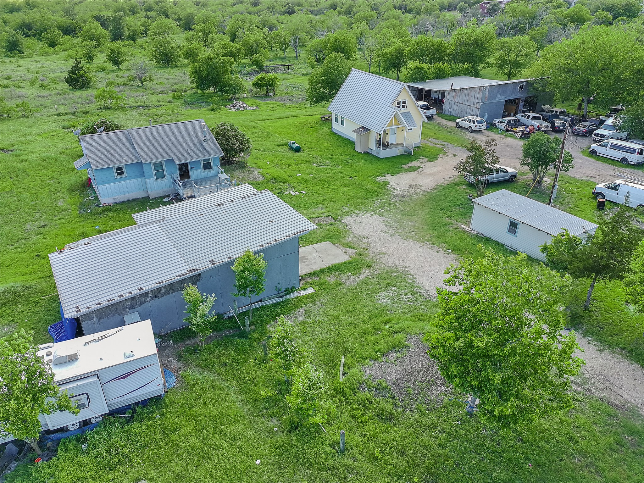 925 Long Hollow Road Dale, TX 78616 - Photo 2 of 26 Multiple structures situated on a verdant landscape, including a light blue residence with a gray shingle roof, a yellow building with a metal roof, and a large gray building with a metal roof