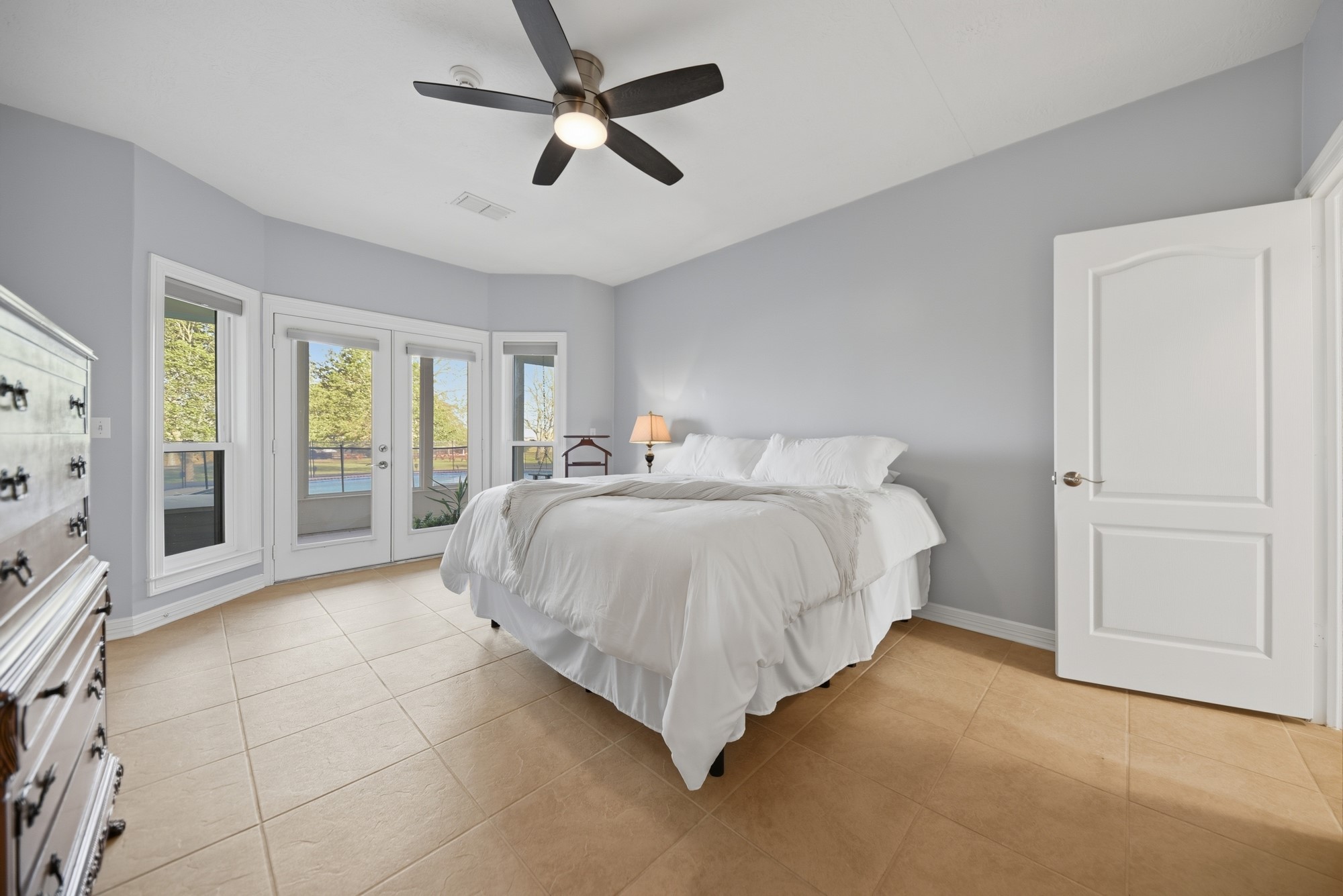 5255 Ganske Road Burton, TX 77835 - Photo 13 of 46 Bright and spacious bedroom with light gray walls and tiled flooring. It features a large bed, ceiling fan, and ample natural light from multiple windows and a glass door leading to an outdoor area.