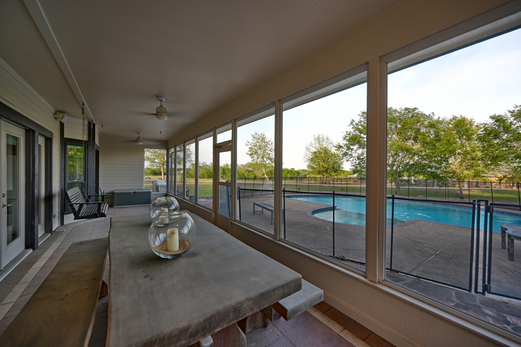5255 Ganske Road Burton, TX 77835 - Photo 15 of 46 Screened-in porch with a large dining table and a swing, overlooking a fenced pool area surrounded by trees, providing a serene outdoor living space.