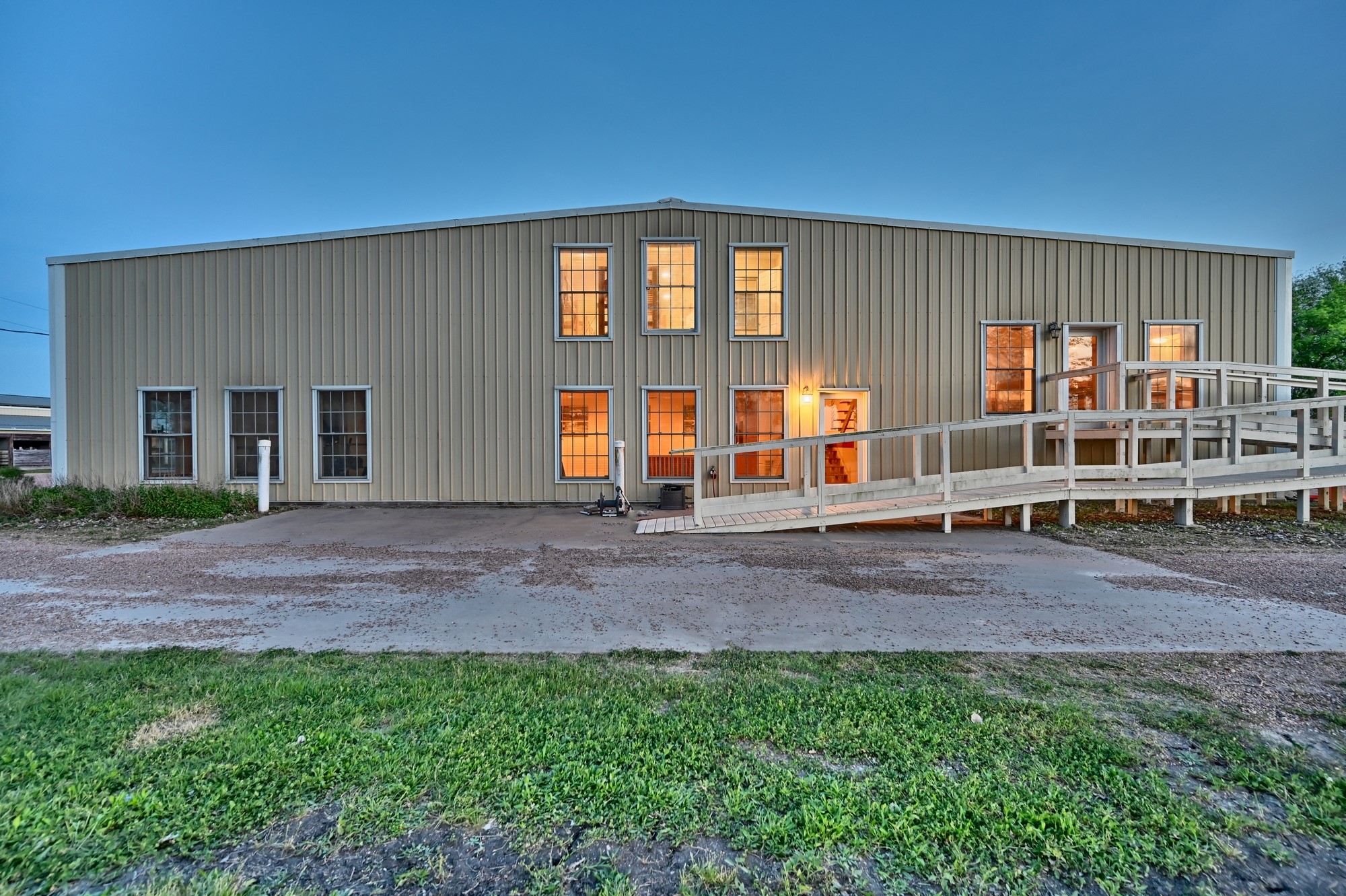 5255 Ganske Road Burton, TX 77835 - Photo 17 of 46 Massive shop and bunk house building with multiple windows and a ramp leading to the entrance. The exterior is clad in beige siding, and the building is surrounded by a gravel driveway and grassy area. The warm interior lighting creates a welcoming atmosphere.