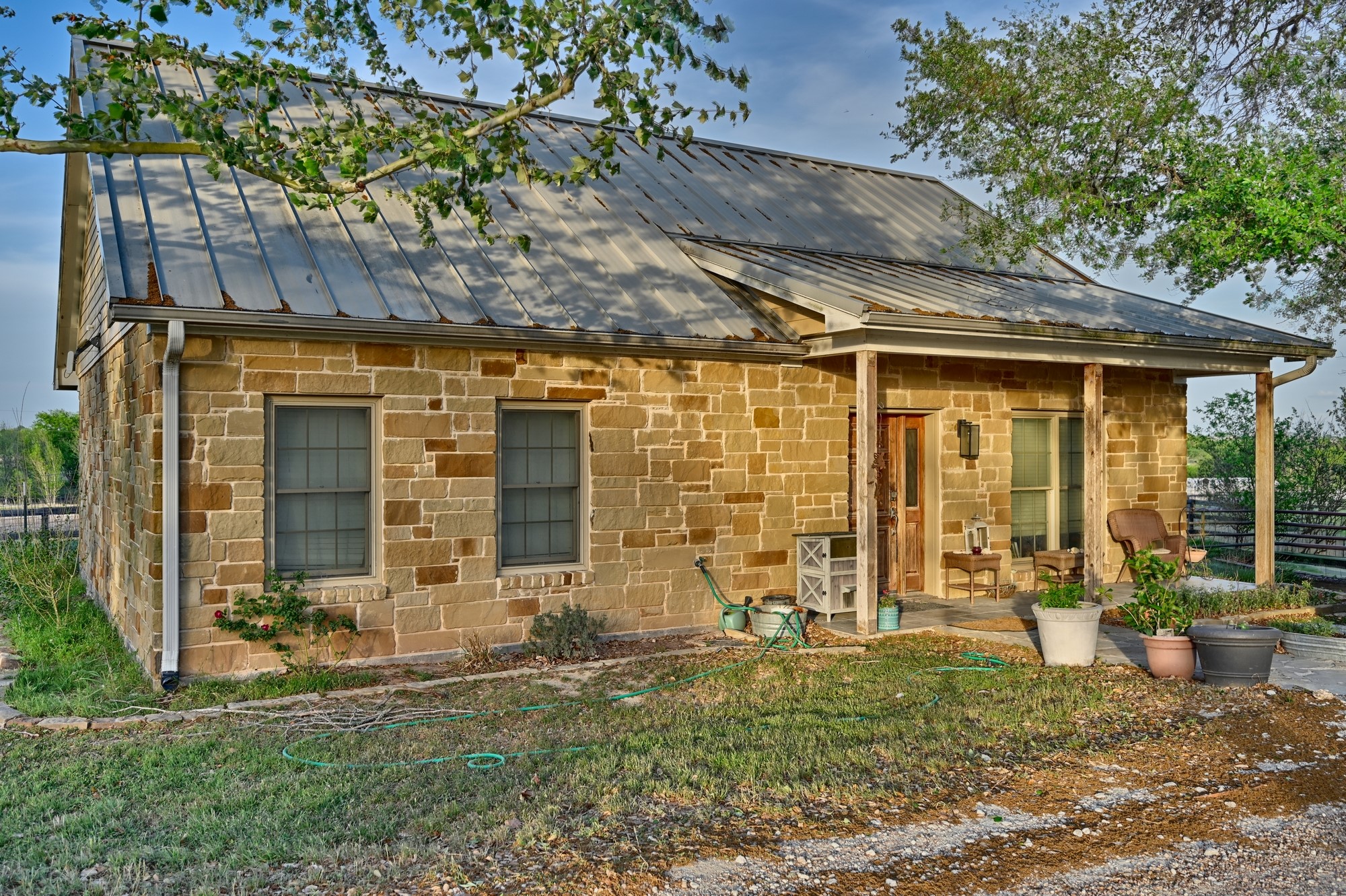 5255 Ganske Road Burton, TX 77835 - Photo 23 of 46 Charming rustic home with stone exterior and metal roof. Features a cozy porch with potted plants, surrounded by lush greenery. Ideal for those seeking a serene, countryside vibe.