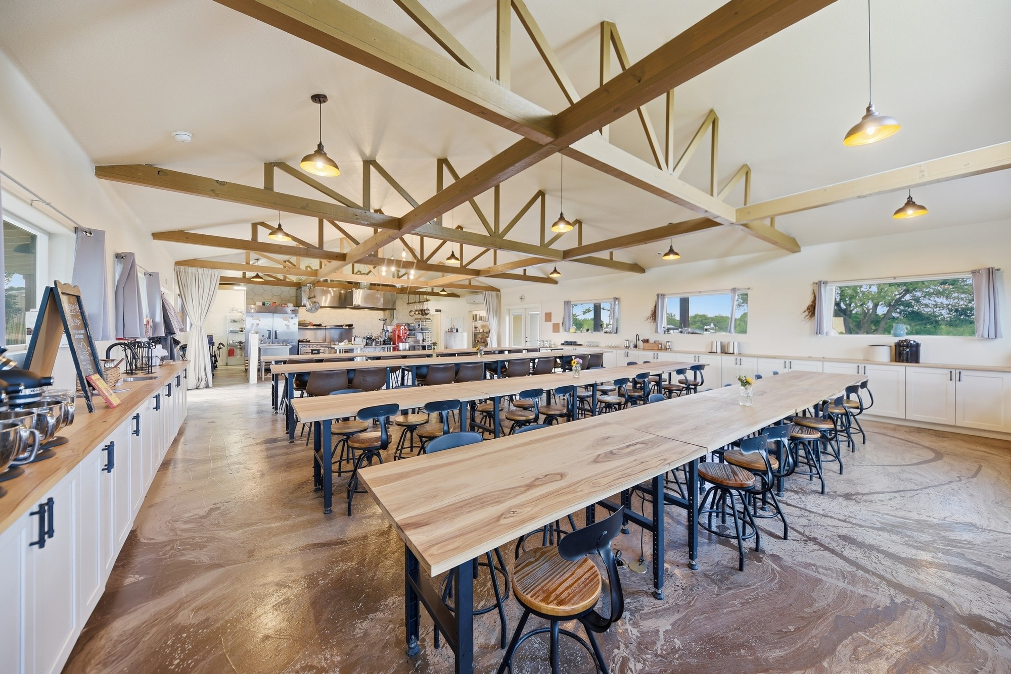 5255 Ganske Road Burton, TX 77835 - Photo 28 of 46 Spacious open-concept kitchen and dining area with high ceilings, exposed wooden beams, and ample natural light. Features long wooden tables, modern lighting fixtures, and extensive counter space, ideal for entertaining or communal dining.
