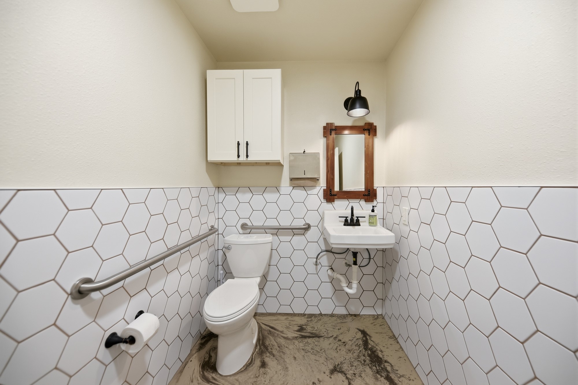5255 Ganske Road Burton, TX 77835 - Photo 30 of 46 2 identical bathrooms features a modern design with hexagonal white tiles covering the lower half of the walls. It includes a compact sink with a stylish black faucet, a wall-mounted mirror, and a practical overhead cabinet for storage. The room is equipped with accessibility rails and has a sleek, minimalist light fixture.
