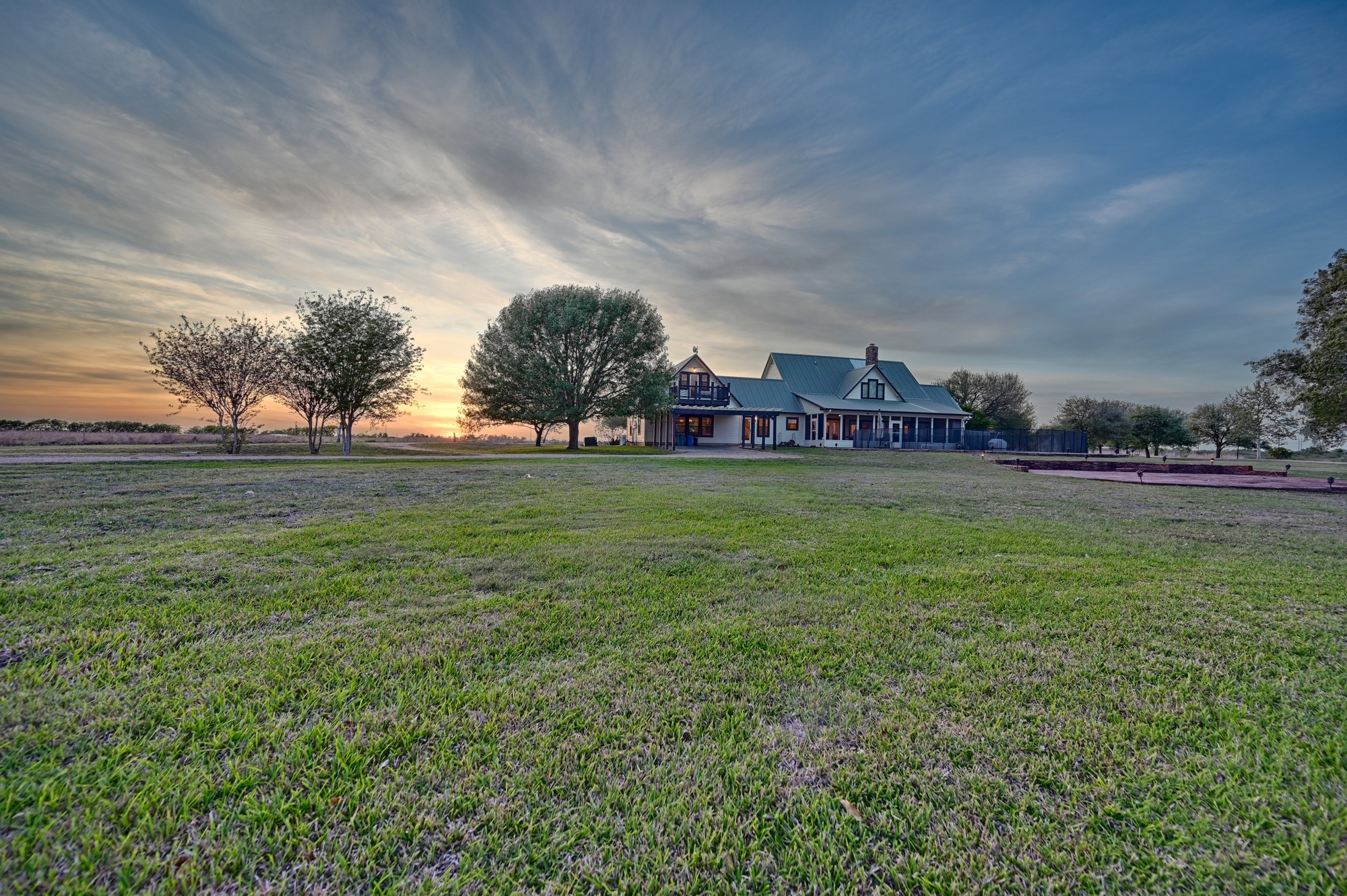 5255 Ganske Road Burton, TX 77835 - Photo 44 of 46 This photo showcases a charming house with a large, open yard, surrounded by mature trees. The sky is painted with a beautiful sunset, adding warmth to the serene rural setting.