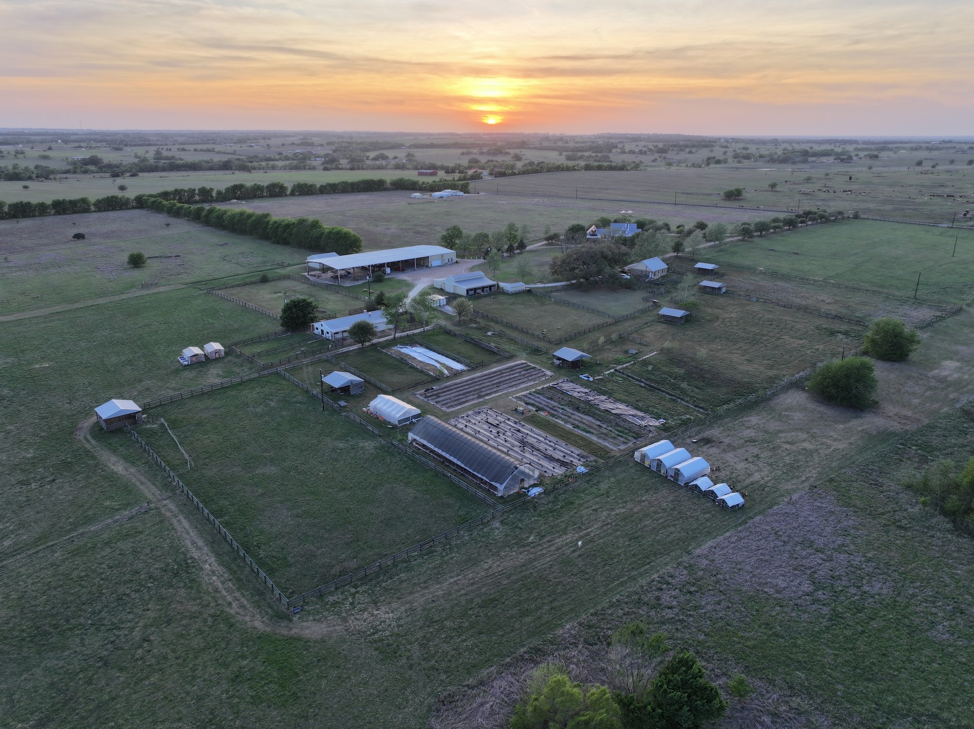 5255 Ganske Road Burton, TX 77835 - Photo 46 of 46 Expansive rural property with multiple structures, including barns and greenhouses, set against a picturesque sunset. Ideal for farming or a serene countryside lifestyle.