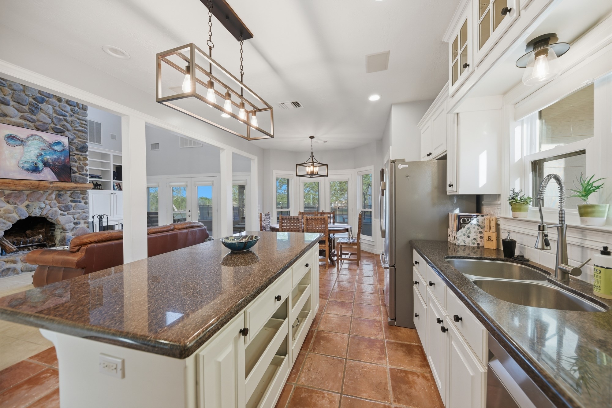 5255 Ganske Road Burton, TX 77835 - Photo 8 of 46 Bright, open kitchen and living area. The kitchen features a large island with granite countertops, modern light fixtures, and stainless steel appliances. Adjacent is a cozy living space with a stone fireplace and large windows offering ample natural light. The dining area is nestled by the windows, creating a welcoming atmosphere.