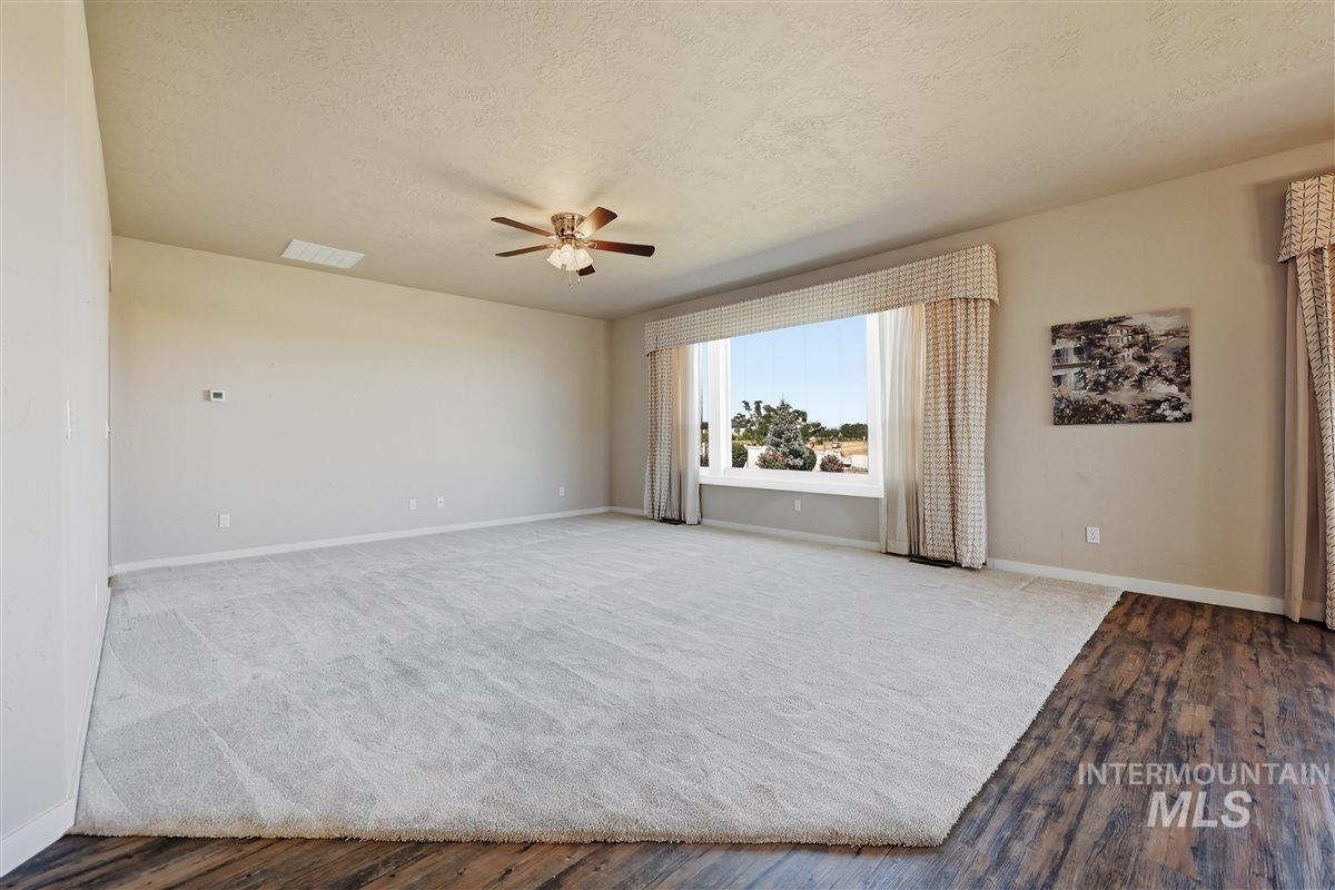 13496 Hockberger Ranch Road Caldwell, ID 83607 - Photo 18 of 40 Spacious room featuring a large picture window with patterned drapes, a ceiling fan, plush grey carpeting, and wood-finish flooring