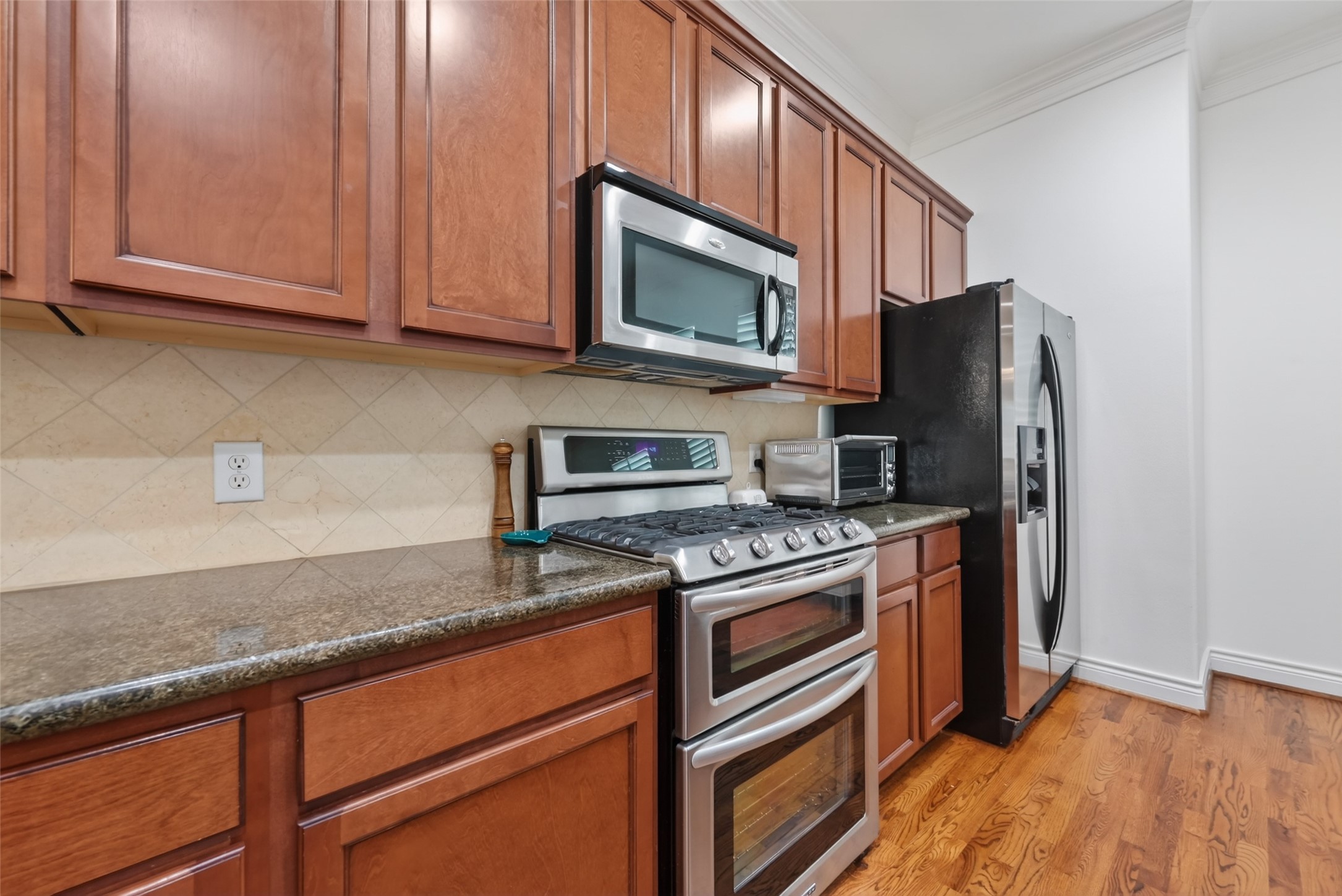 620 Rutland Street, Unit A Houston, TX 77007 - Photo 17 of 46 This kitchen features warm wood cabinetry, granite countertops, and stainless steel appliances, including a double oven and side-by-side refrigerator. The hardwood floors add a touch of elegance, while the neutral backsplash provides a clean, classic look.