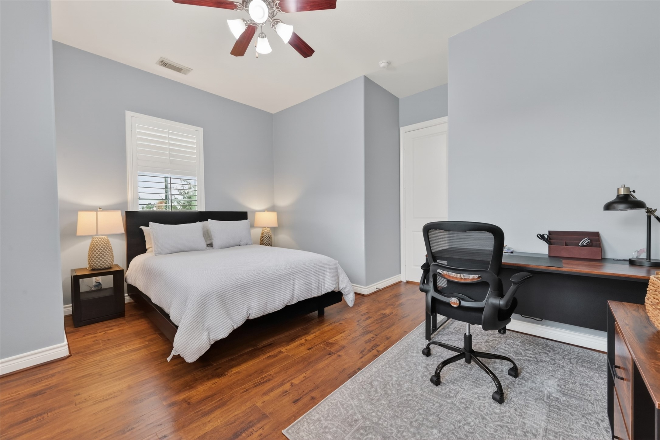 620 Rutland Street, Unit A Houston, TX 77007 - Photo 21 of 46 This bedroom features a comfortable bed, wooden flooring, and soft gray walls. It includes a ceiling fan, ample natural light from a window with shutters, and a cozy workspace with a modern desk and chair.
