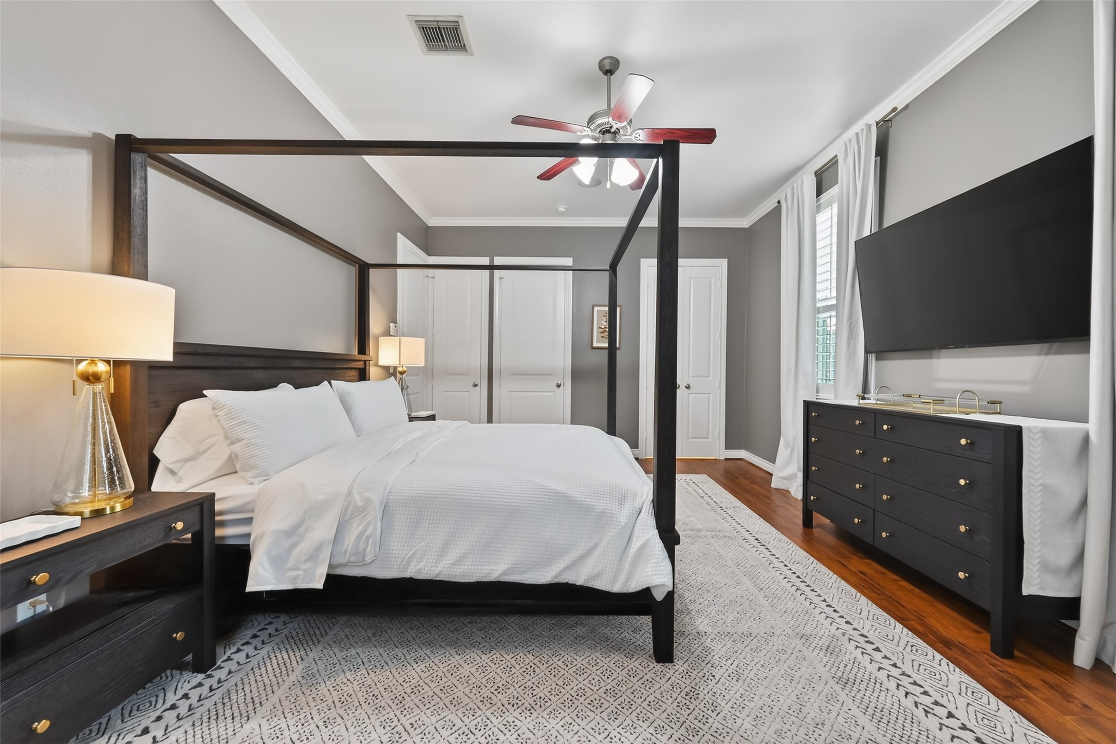 620 Rutland Street, Unit A Houston, TX 77007 - Photo 24 of 46 This bedroom features a stylish four-poster bed with crisp white linens, complemented by a modern dresser and nightstands. The room is accented with a ceiling fan, large TV, and floor-to-ceiling windows for natural light. Elegant gray walls and a patterned rug complete the sophisticated look.